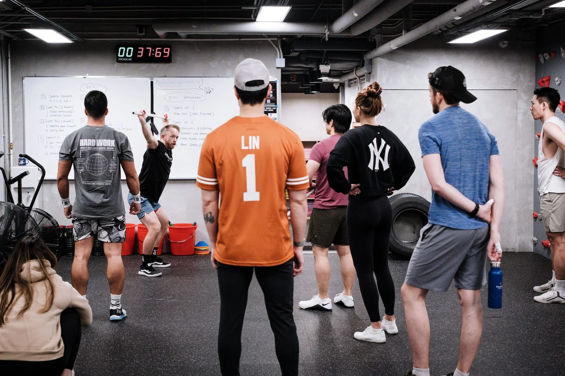 Group of people participating in a fitness class or workshop in a gym, with a trainer leading the session at the whiteboard, and a digital timer above showing 00:37:69.