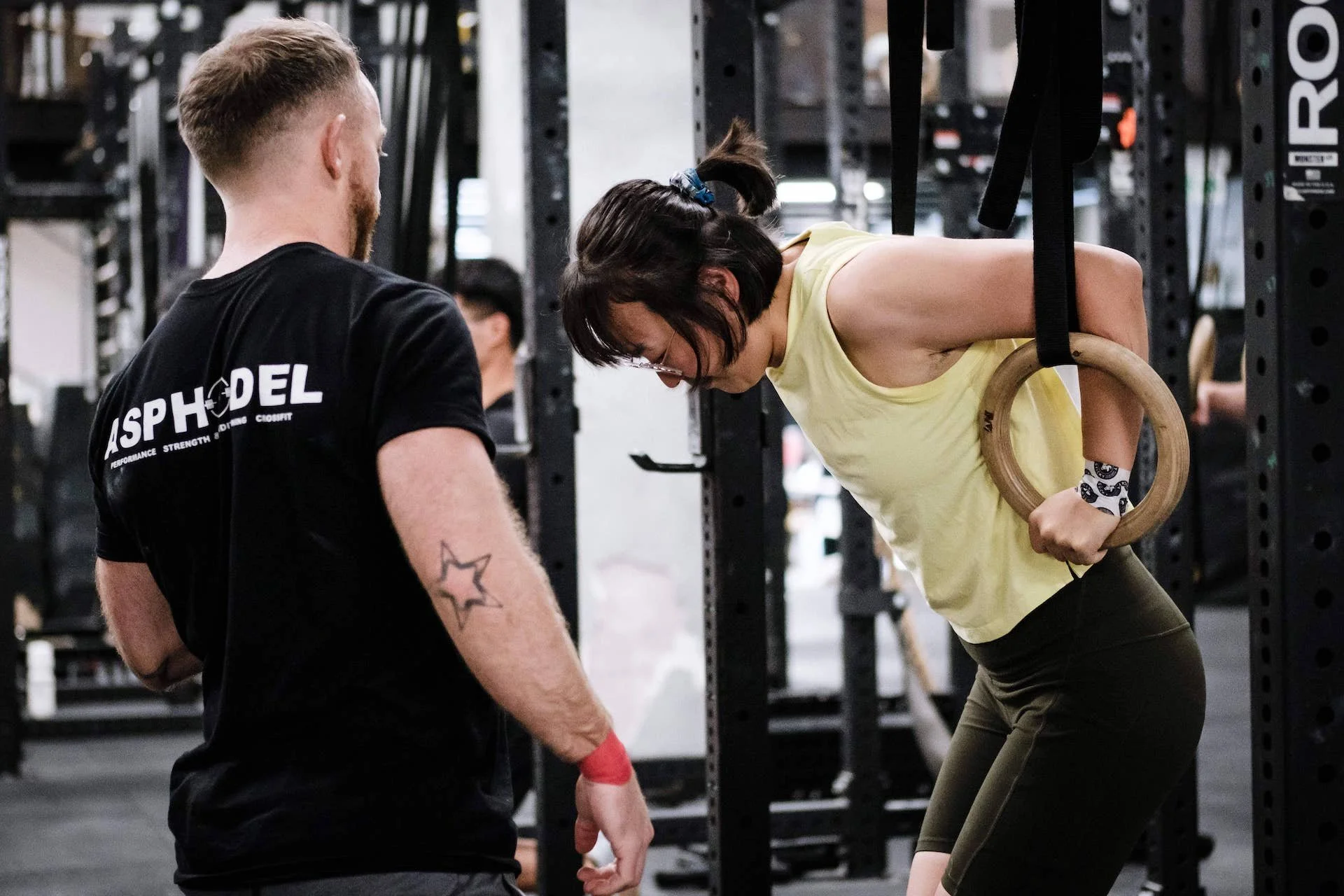 A woman in a yellow tank top and black leggings performs an exercise using gymnastic rings held overhead, assisted by a trainer in a black t-shirt at a gym.