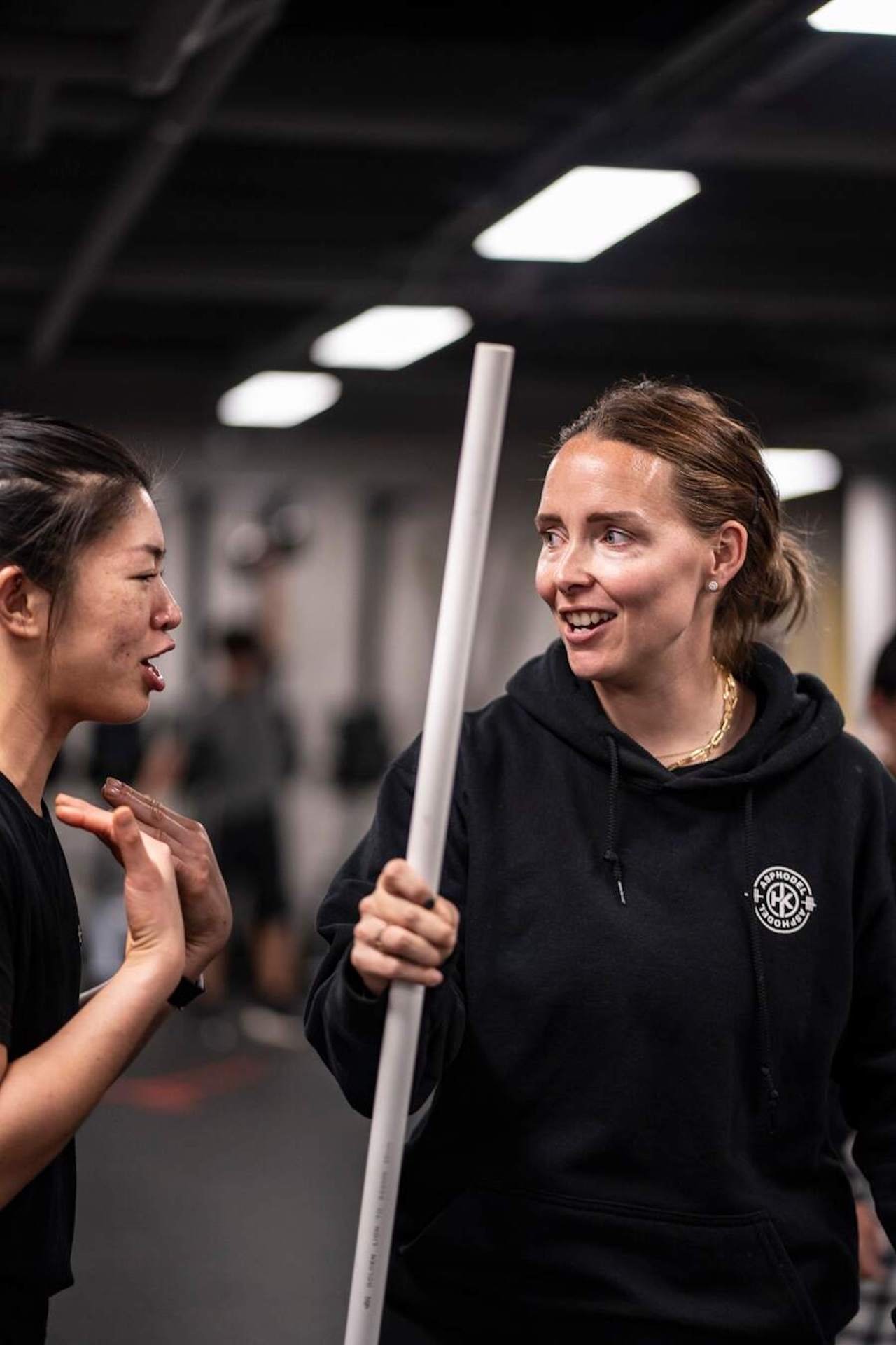 Two women in a gym having a conversation. One woman is holding a white pole, and the other woman appears to be actively gesturing while talking.