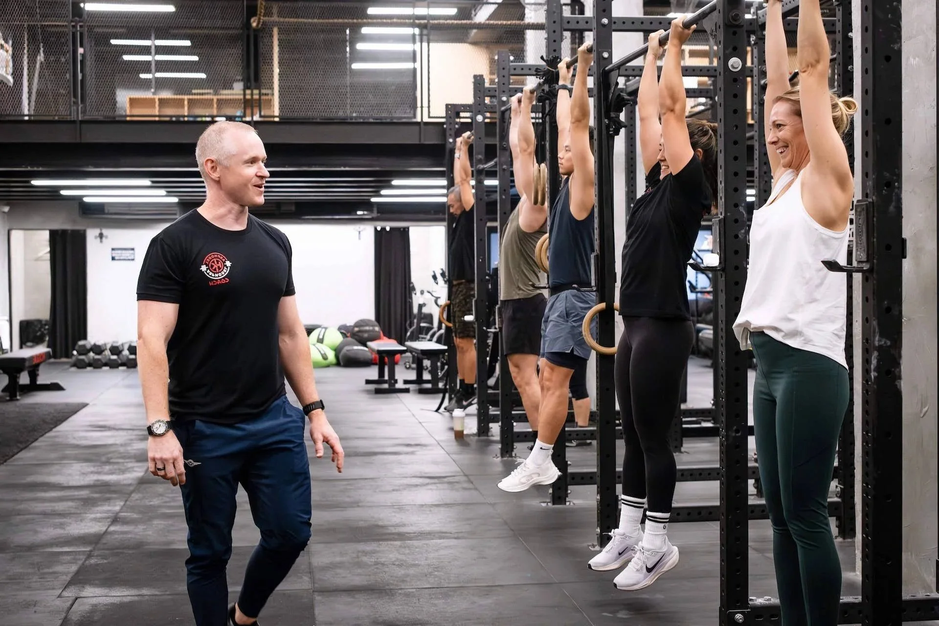 A group of women doing pull-ups in a gym while a male trainer observes and talks to them.