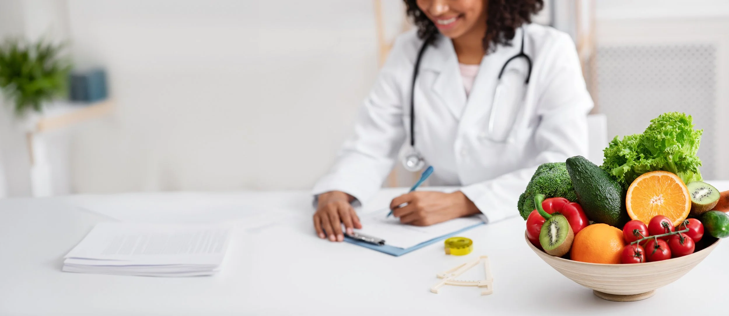 A female doctor in a white coat smiling and writing on a clipboard at a desk, with fresh vegetables and fruits in a bowl in the foreground.
