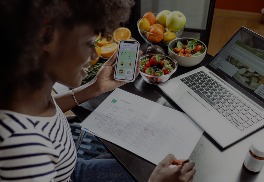 A woman sitting at a table with a laptop, smartphone, planner, and bowls of fruit, including apples, oranges, strawberries, and cherry tomatoes.