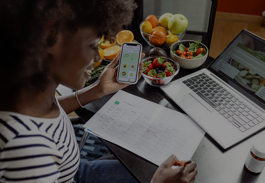 A woman looking at her phone with a diet app, sitting at a table with a laptop, a grocery list, bowls of fresh fruits and vegetables.