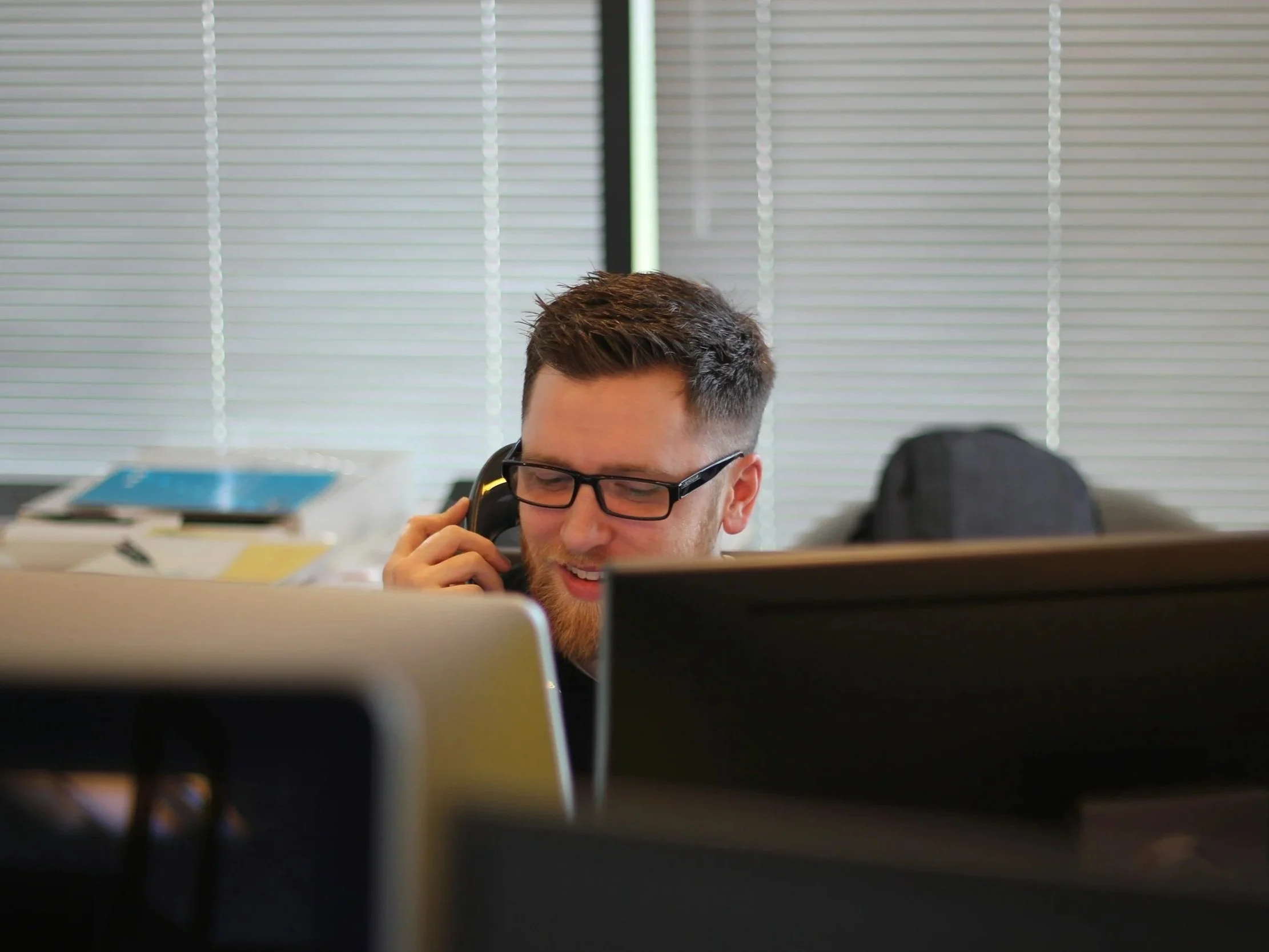 A young man with glasses and a beard talking on the phone in an office setting with stacked papers and files in the background.