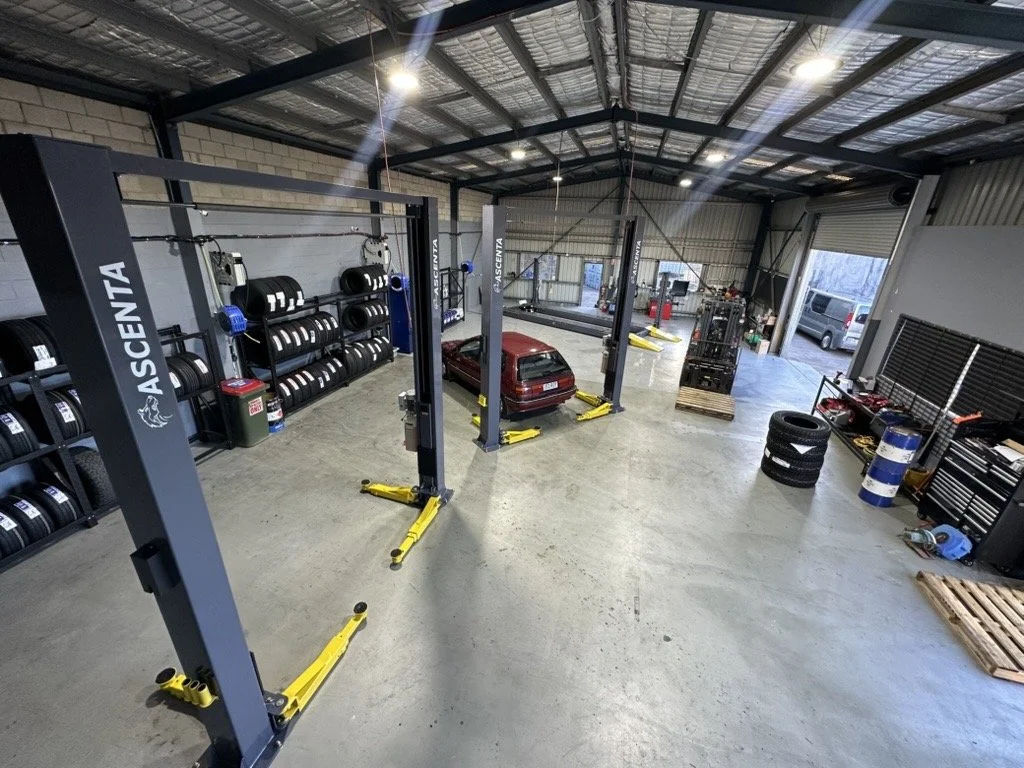 Inside an auto repair shop or garage with vehicle lifts, car tires, tools, and equipment visible.