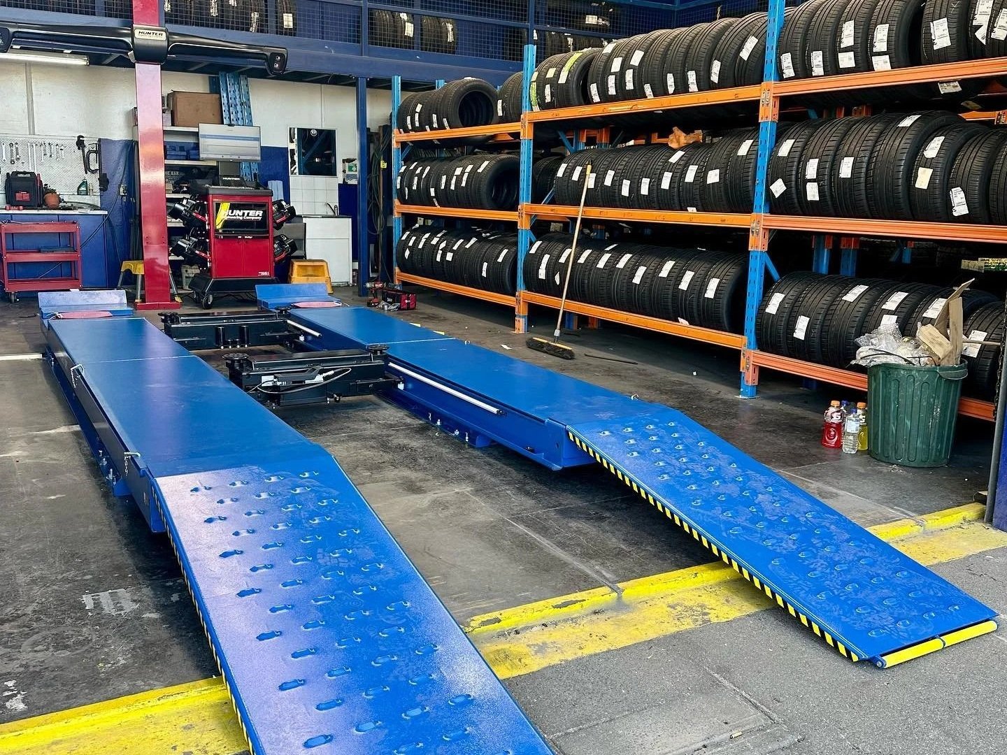 A tire shop with numerous stacks of tires on orange and blue shelves. In the foreground, a blue vehicle lift with ramps is positioned on a concrete floor. Tools, a red rolling tool chest, and a trash bin are visible in the background.