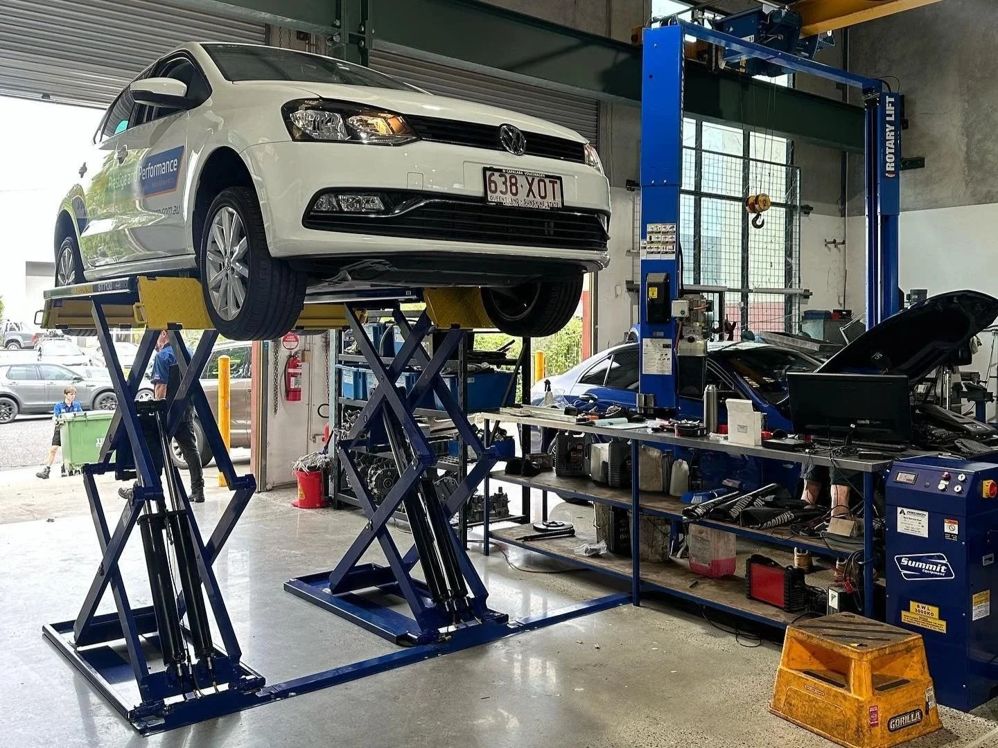 A white car is elevated on a hydraulic lift inside a garage or auto repair shop, with another car and mechanic working in the background, tools and equipment are on the workbench.