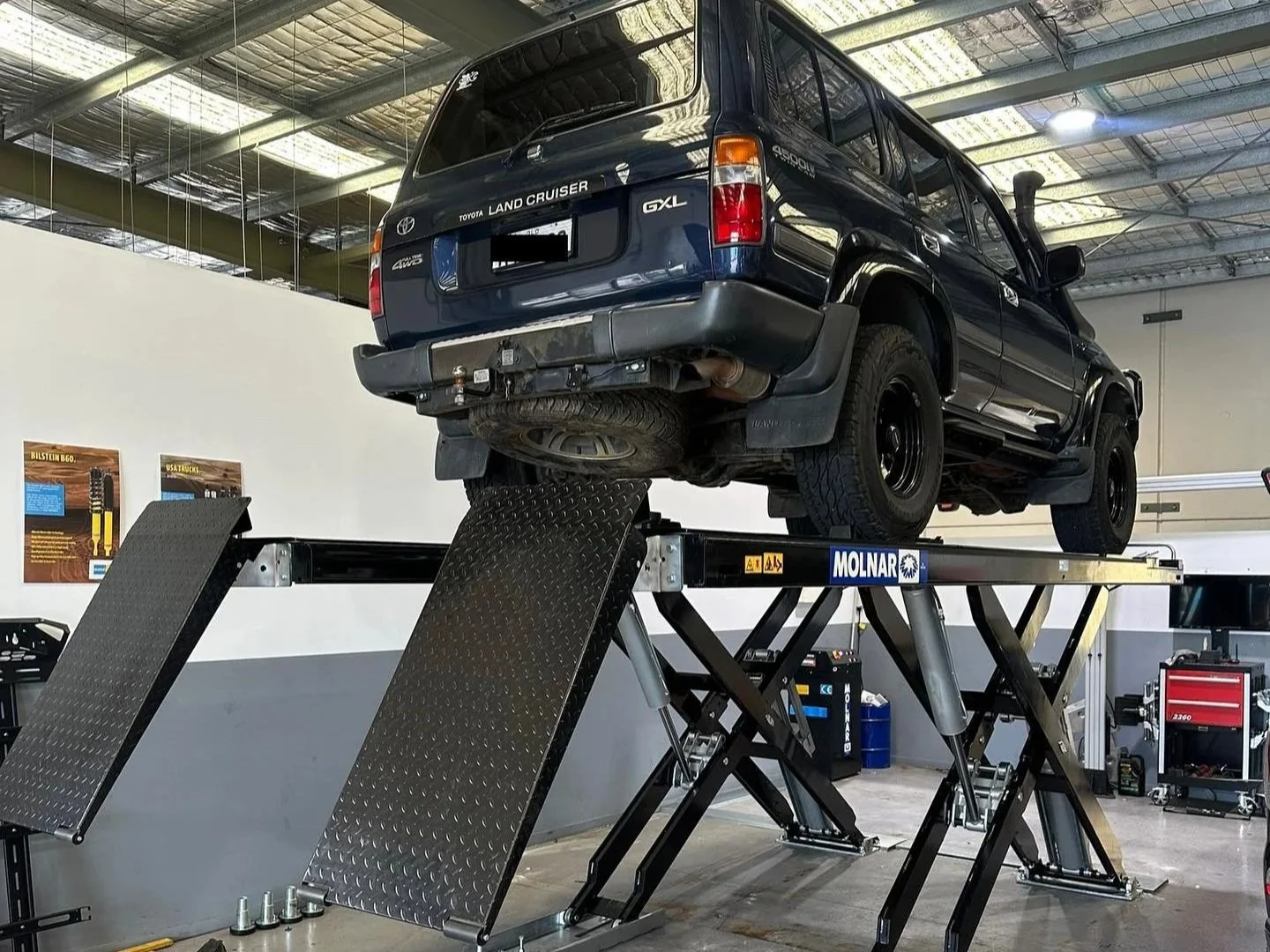 A black Toyota Land Cruiser GXL is elevated on a hydraulic lift inside an auto repair shop.
