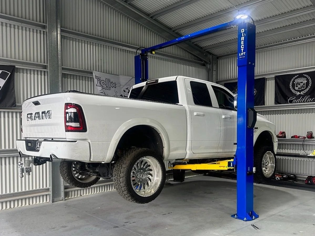 White RAM pickup truck elevated on a blue automotive lift inside a metal-walled garage.