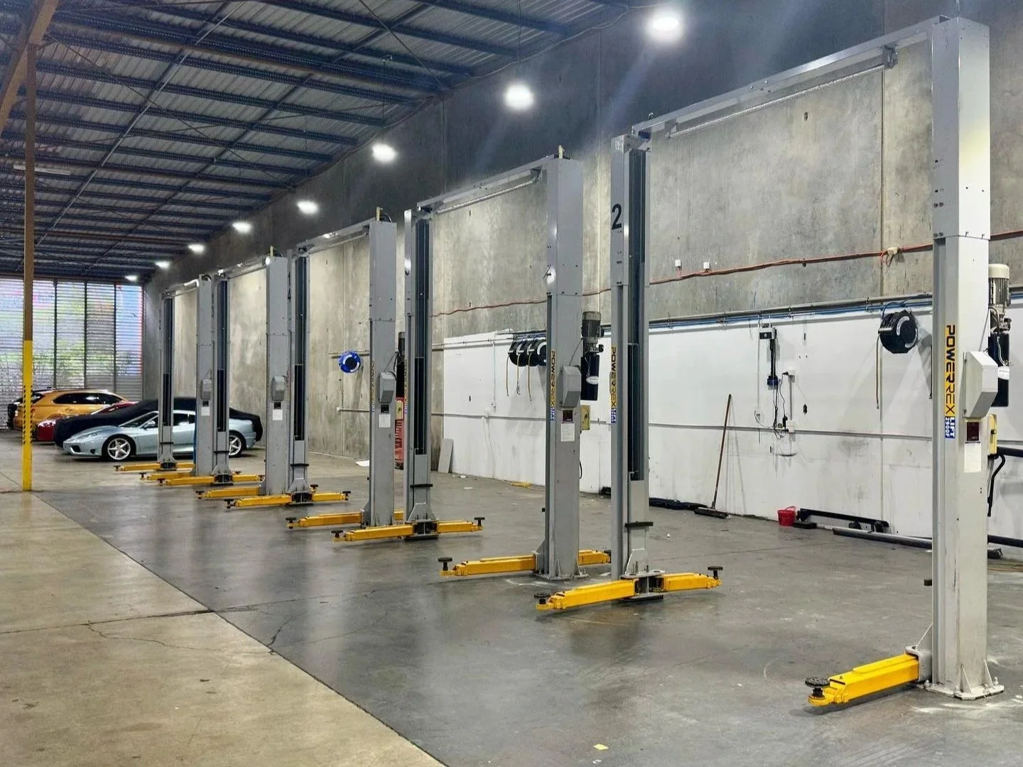 Indoor automotive workshop with five vehicle lifts and several luxury sports cars parked on the left side.