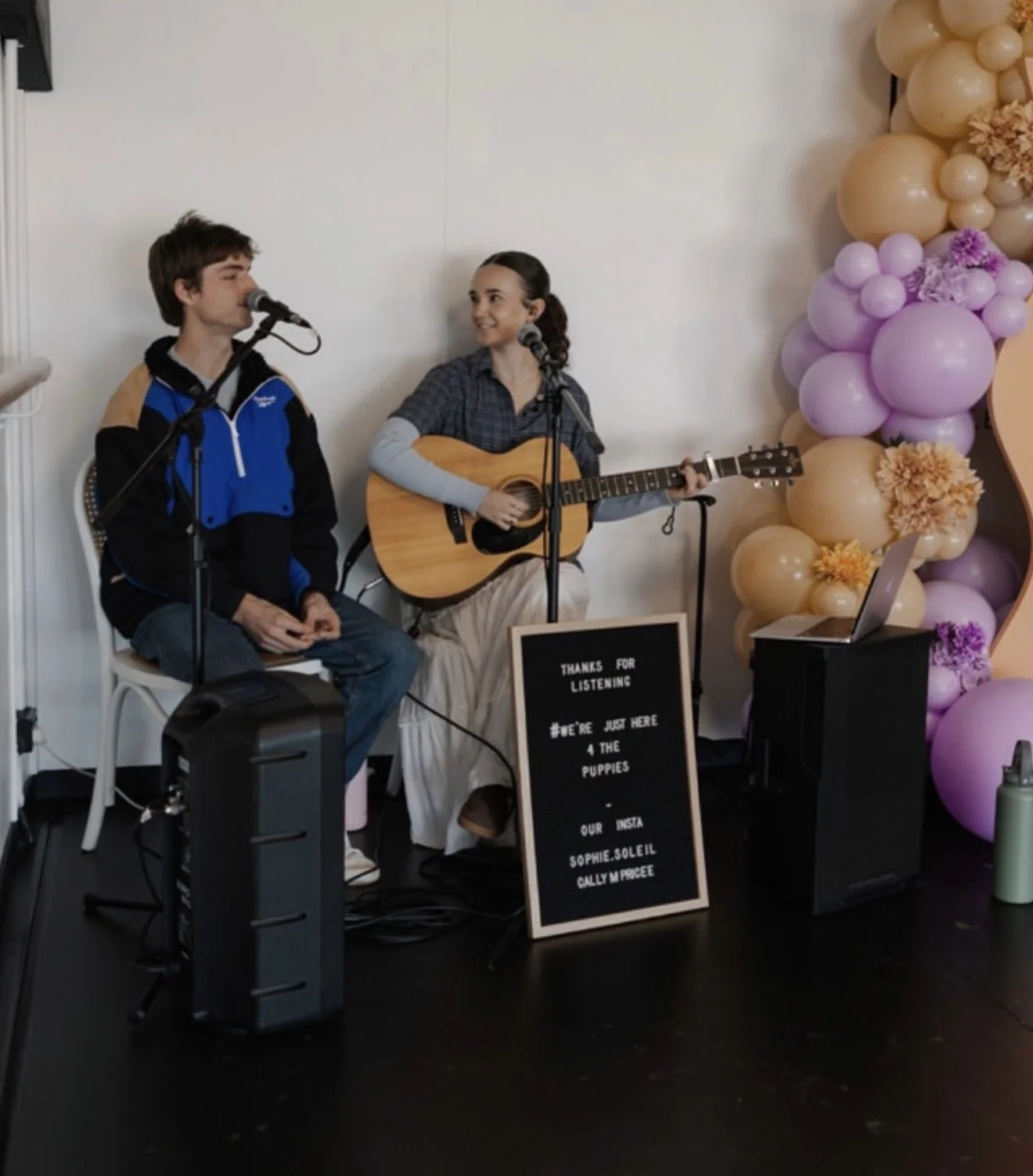 Two teenagers performing music on stage; girl playing guitar and singing, boy holding a microphone; decorative balloons in pastel colors in background; sign with event details in front.