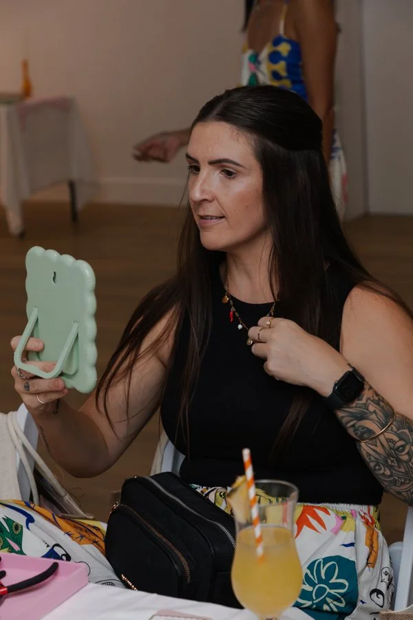 A woman with long dark hair and a tattoo on her arm is holding a mint green phone with a pop socket, taking a selfie at a party or gathering. She is wearing a black sleeveless top, a colorful necklace, and a smartwatch. There are drinks and a black p