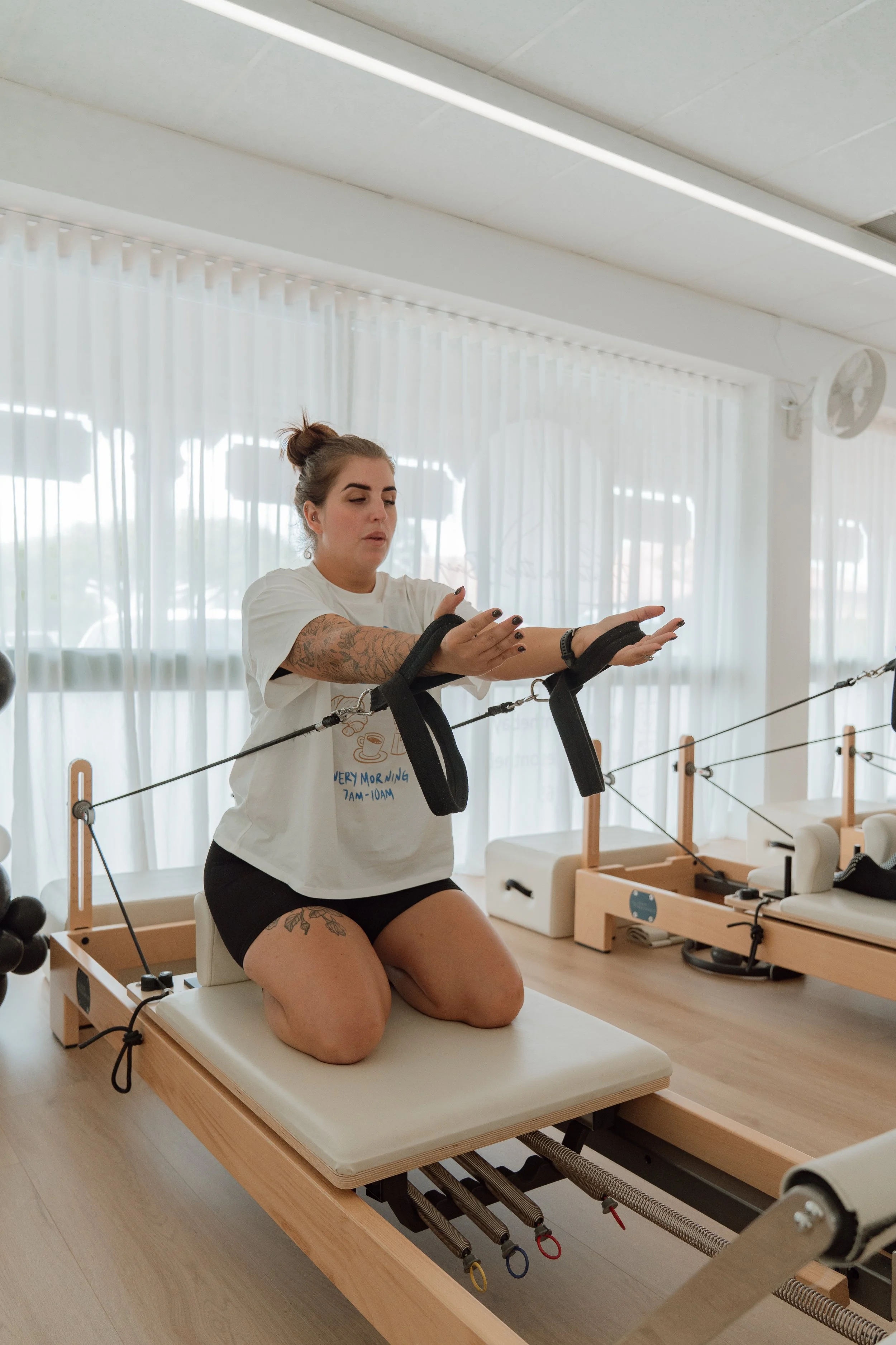 A woman kneeling on a Pilates reformer machine, using black resistance straps with her arms extended forward, in a bright gym with white curtains and multiple Pilates machines.