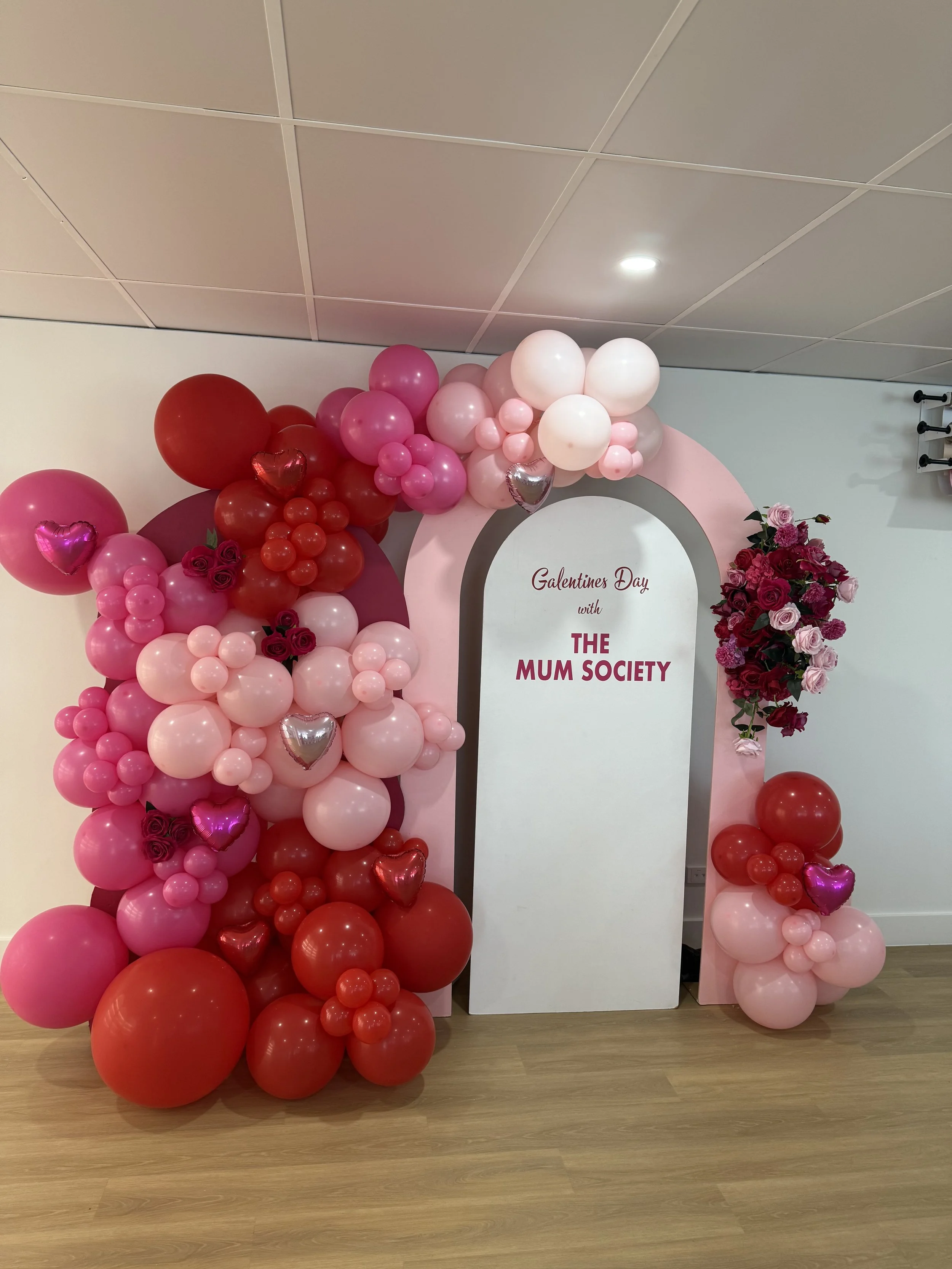 Valentine's Day display with pink, red, and white balloons, and pink and red roses, with a sign that says "Galentines Day with THE MUM SOCIETY".