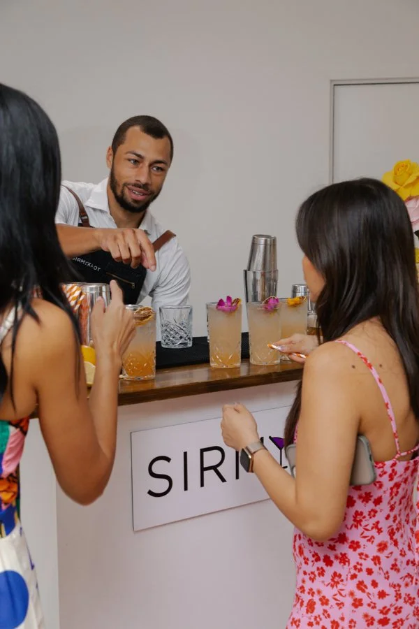 A bartender preparing drinks for two women at a bar with a 'SIR' sign, both women holding drinks and engaging in conversation.