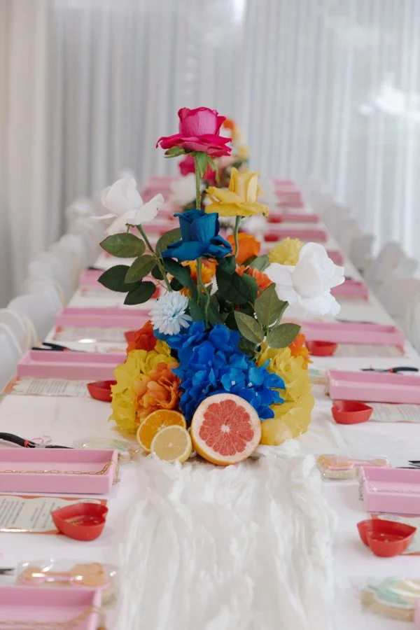 Colorful floral centerpiece with pink, yellow, white, blue, and orange flowers on a long dining table with pink special occasion place cards and red cups, set with utensils and menus, in a bright room with sheer curtains.