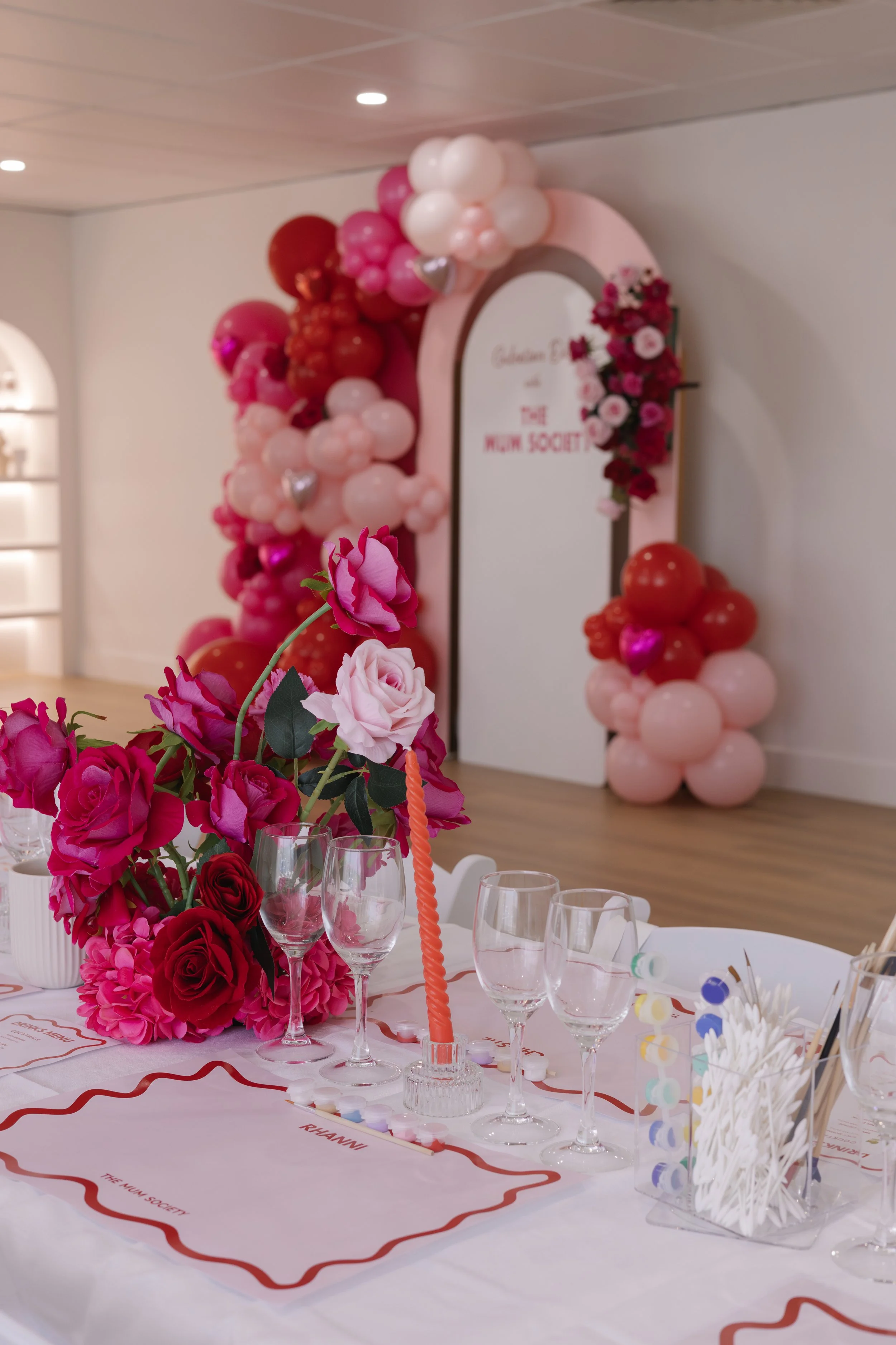 Decorated table with pink and red flowers, empty wine glasses, and a pink balloon arch in the background with a pink and white sign reading "Golden B" and "The Mum Society".