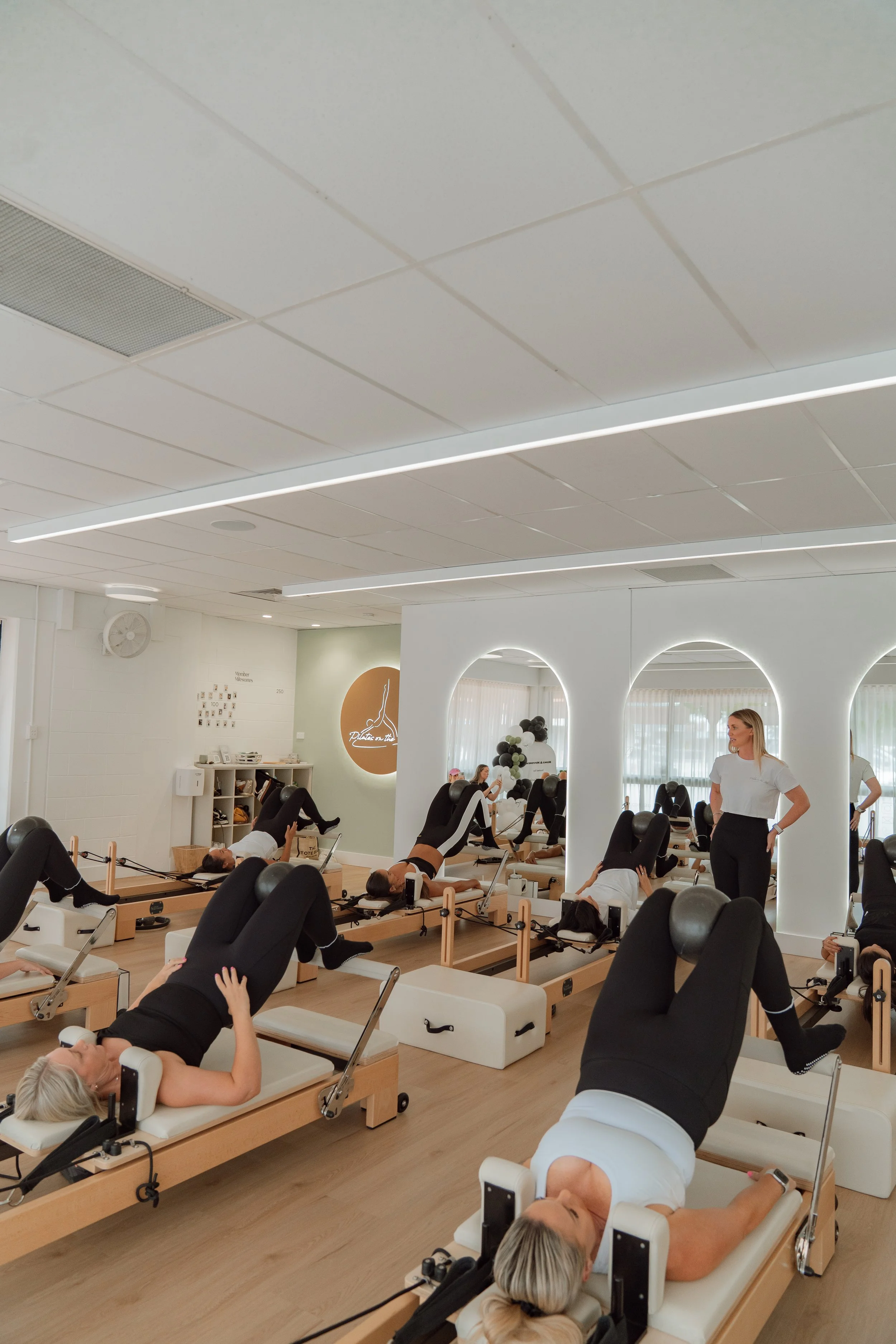 Women in a fitness class using Pilates reformer machines, with an instructor observing, in a modern studio with white walls, mirrors, and natural light.