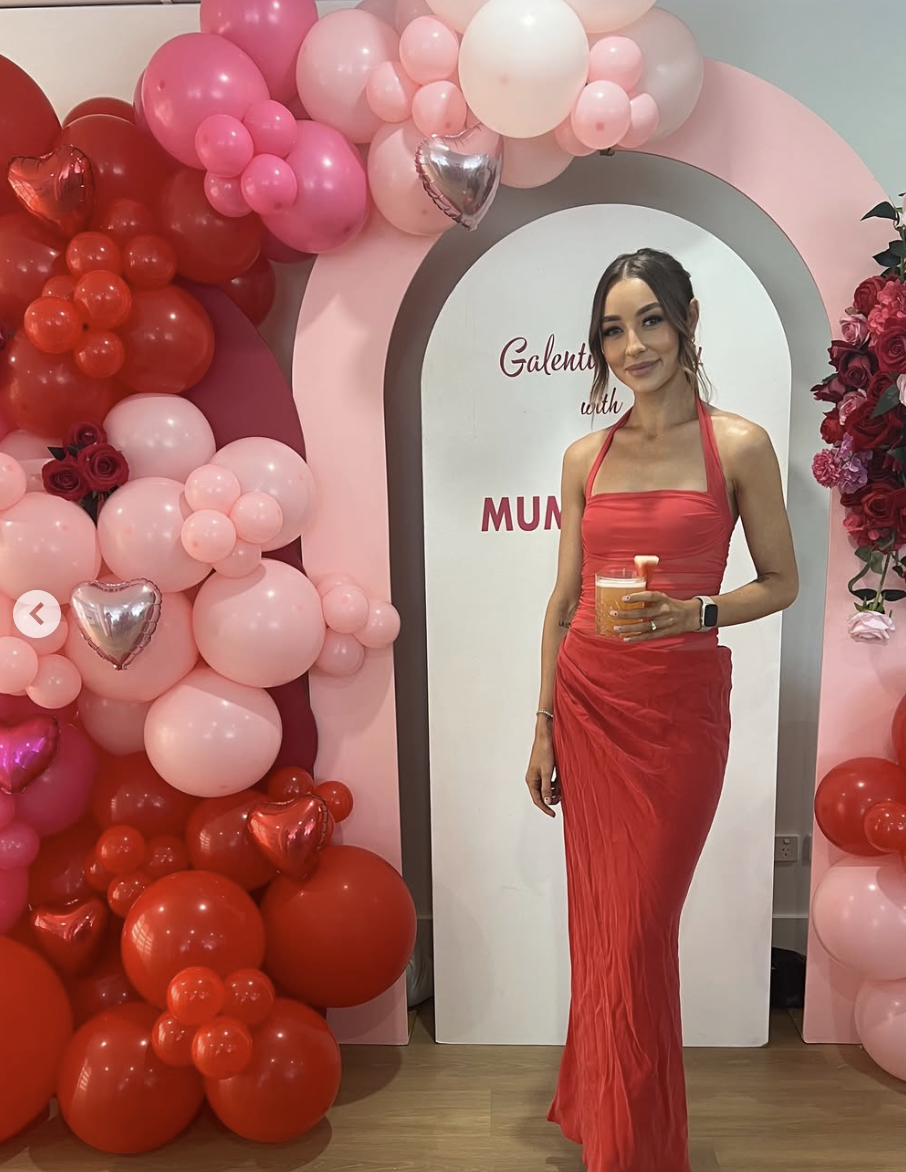 A woman in a red dress standing in front of a balloon arch with pink and red balloons, some heart-shaped, at a celebration or event with a white backdrop that has pink and red text.