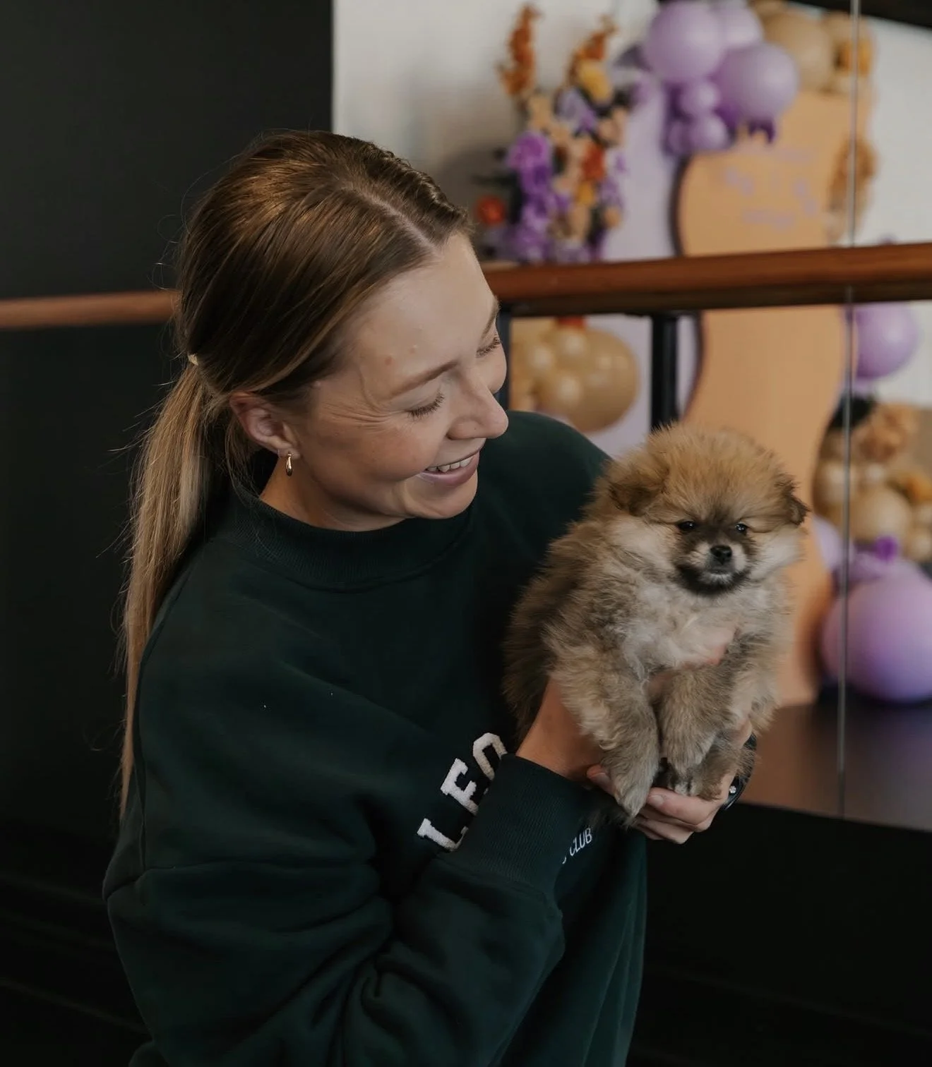 A woman holding a fluffy Pomeranian puppy, smiling happily, in an indoor setting with decorative objects in the background.
