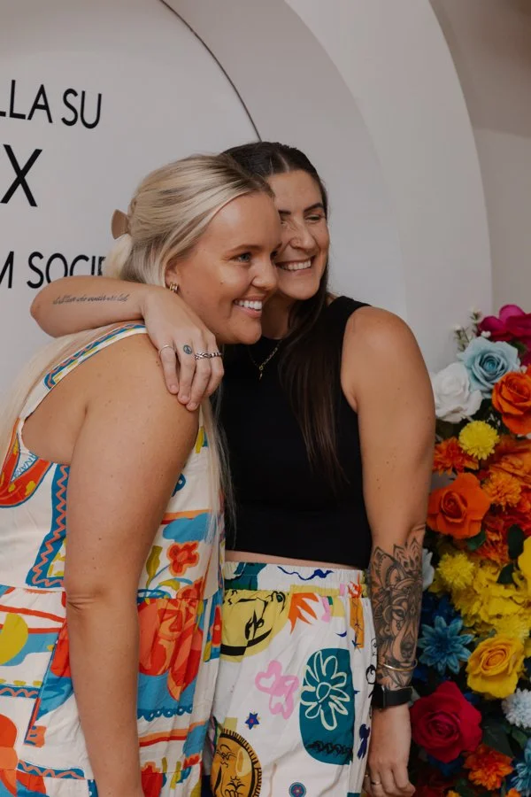Two women hugging and smiling in front of a white wall with text, beside colorful flower arrangements.