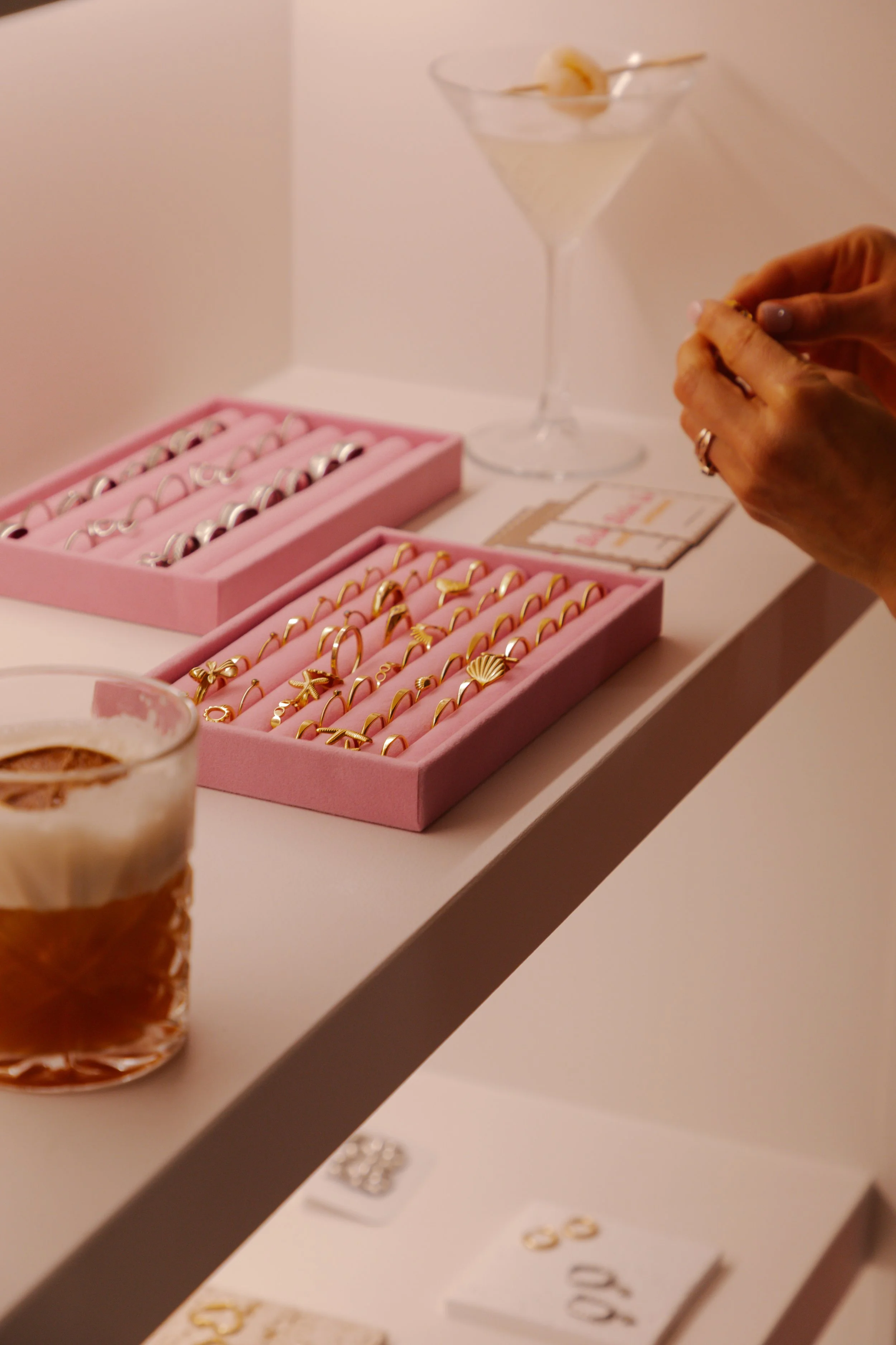 Pink jewelry display tray with gold rings and jewelry, a hand holding a ring, a martini glass with an olive, and a latte in a glass cup on a white table.
