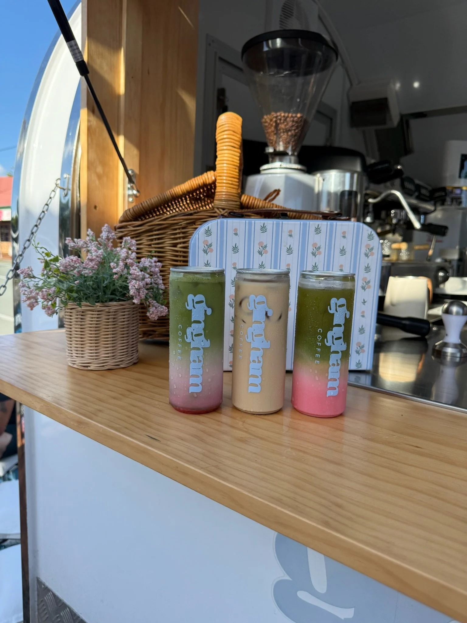 Three colorful smoothies in tall cans with 'Fightram Coffee' printed on them, placed on a wooden counter next to a potted pink flower, with coffee brewing equipment in the background.