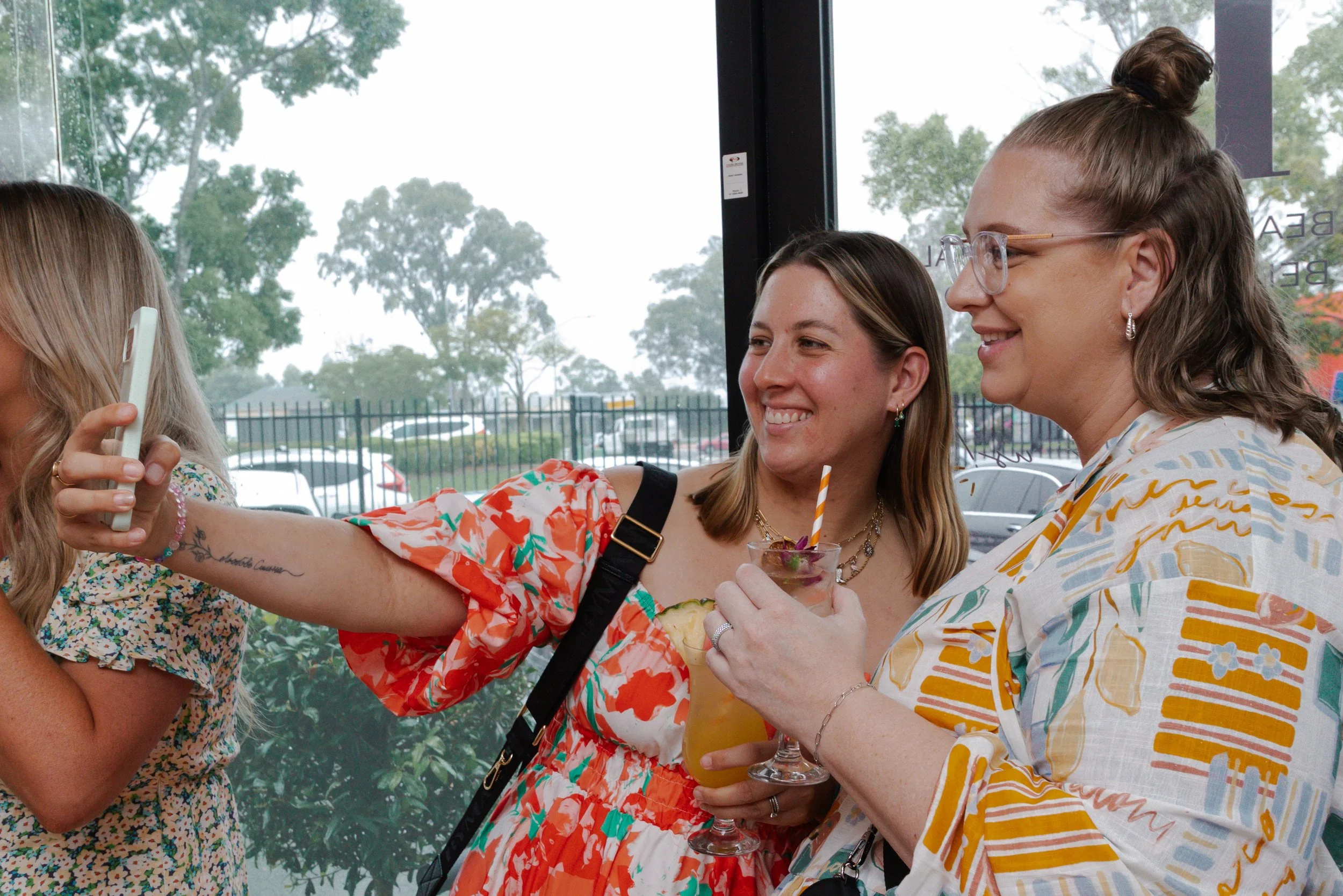 Two women smiling and taking a selfie together inside a cafe, with one holding a colorful drink with a straw.