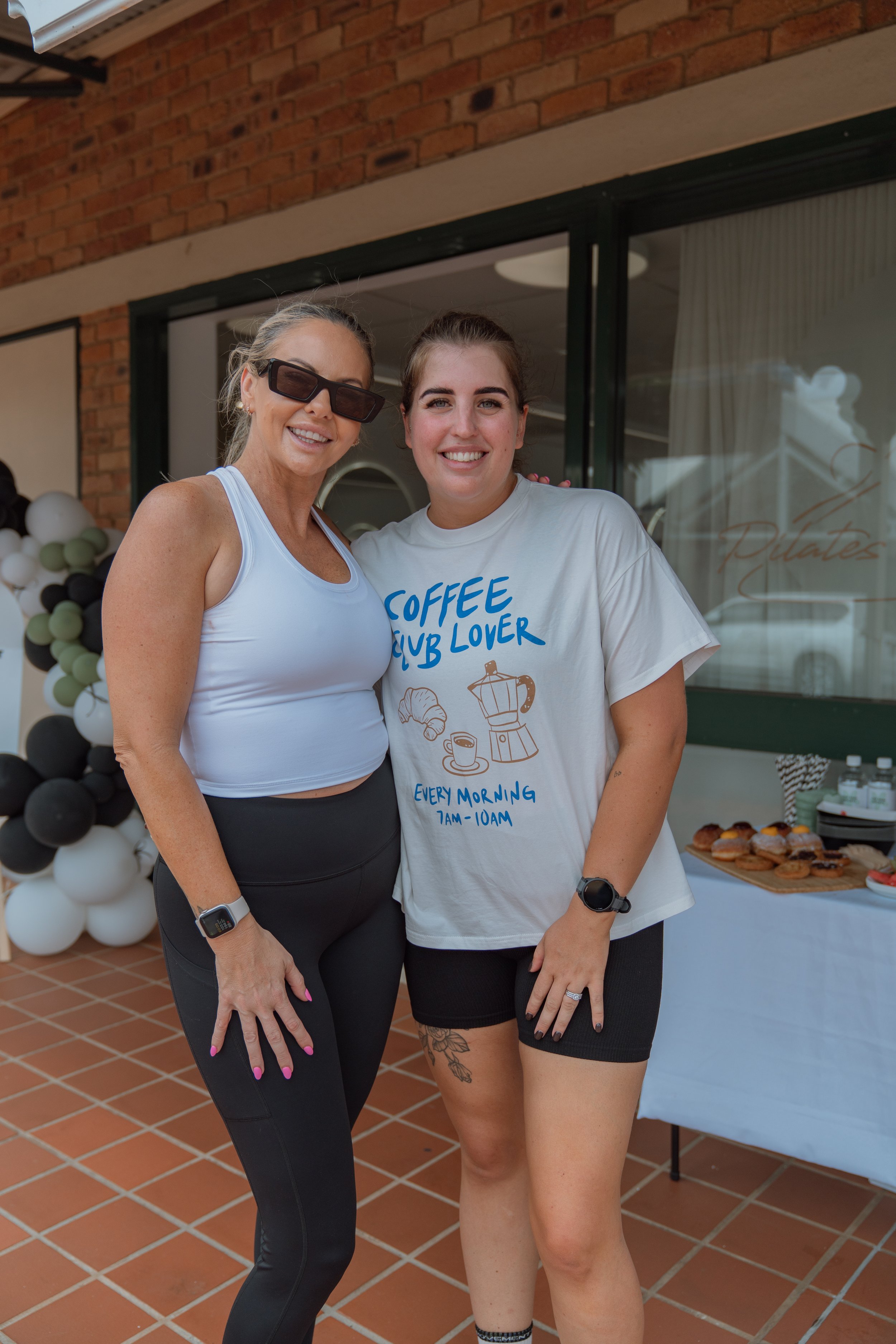 Two women standing outdoors near a brick building, smiling at the camera. One is wearing sunglasses, a white crop top, and black leggings; the other is wearing a white t-shirt with blue text and black shorts. There is a table with food and drinks beh