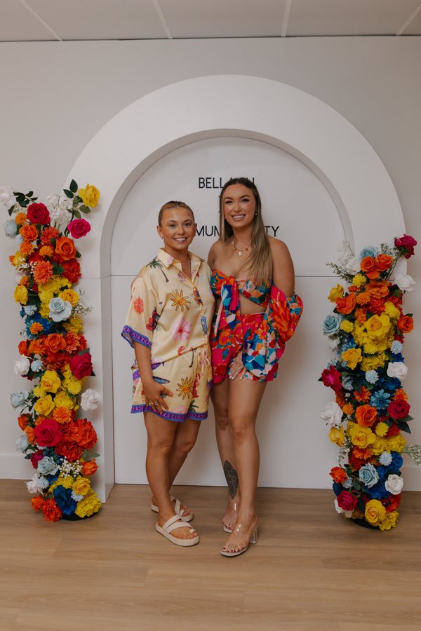 Two women standing under a white decorative arch framed by colorful floral arrangements, smiling at the camera.