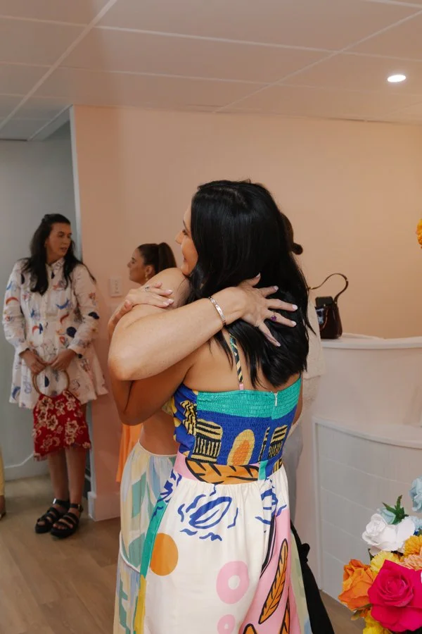 Two women hugging at a social gathering, with other women in the background and colorful flowers in the foreground.