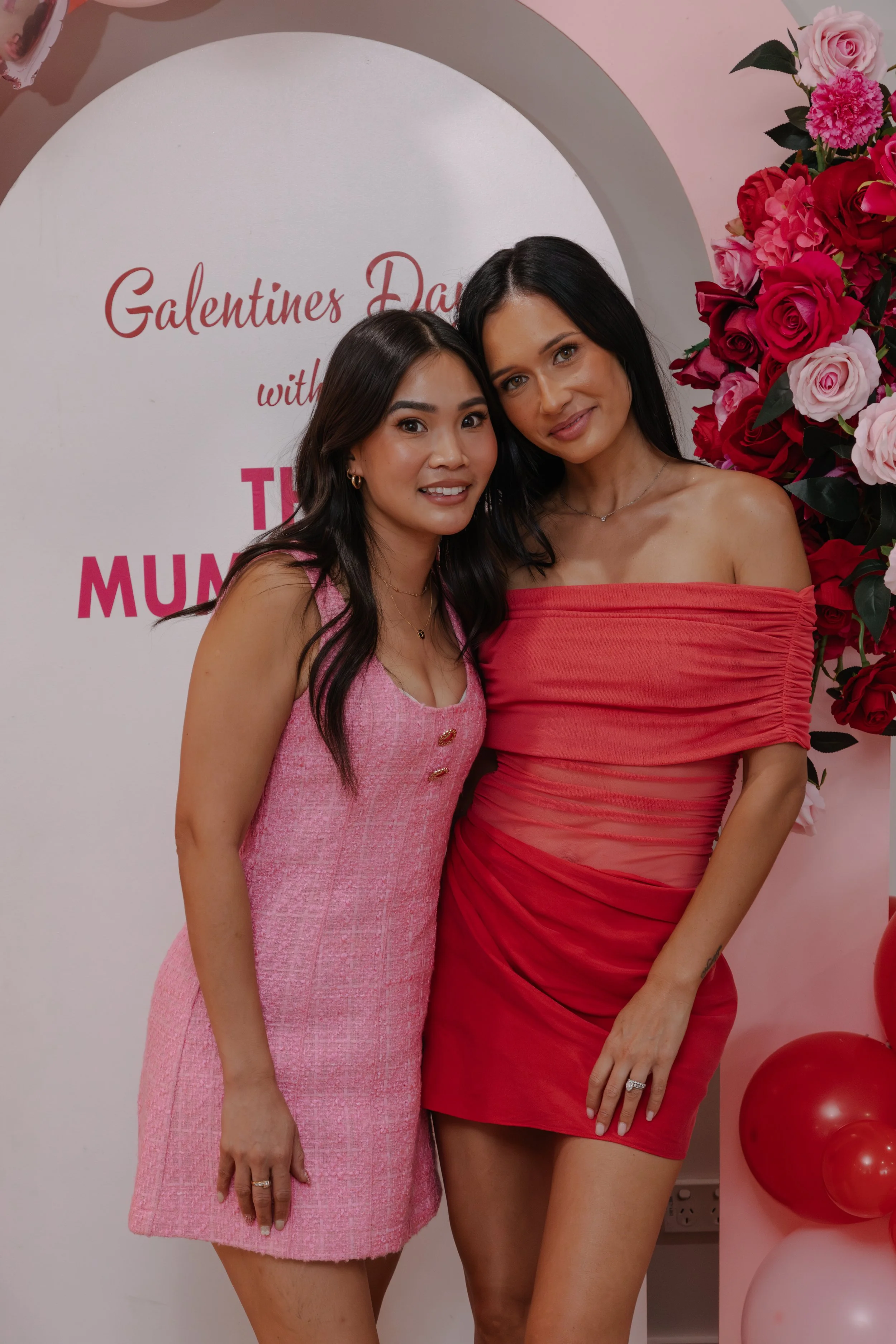 Two women posing together at a Valentine's Day event, with a pink and red floral backdrop and a sign that reads 'Galentines Day'.