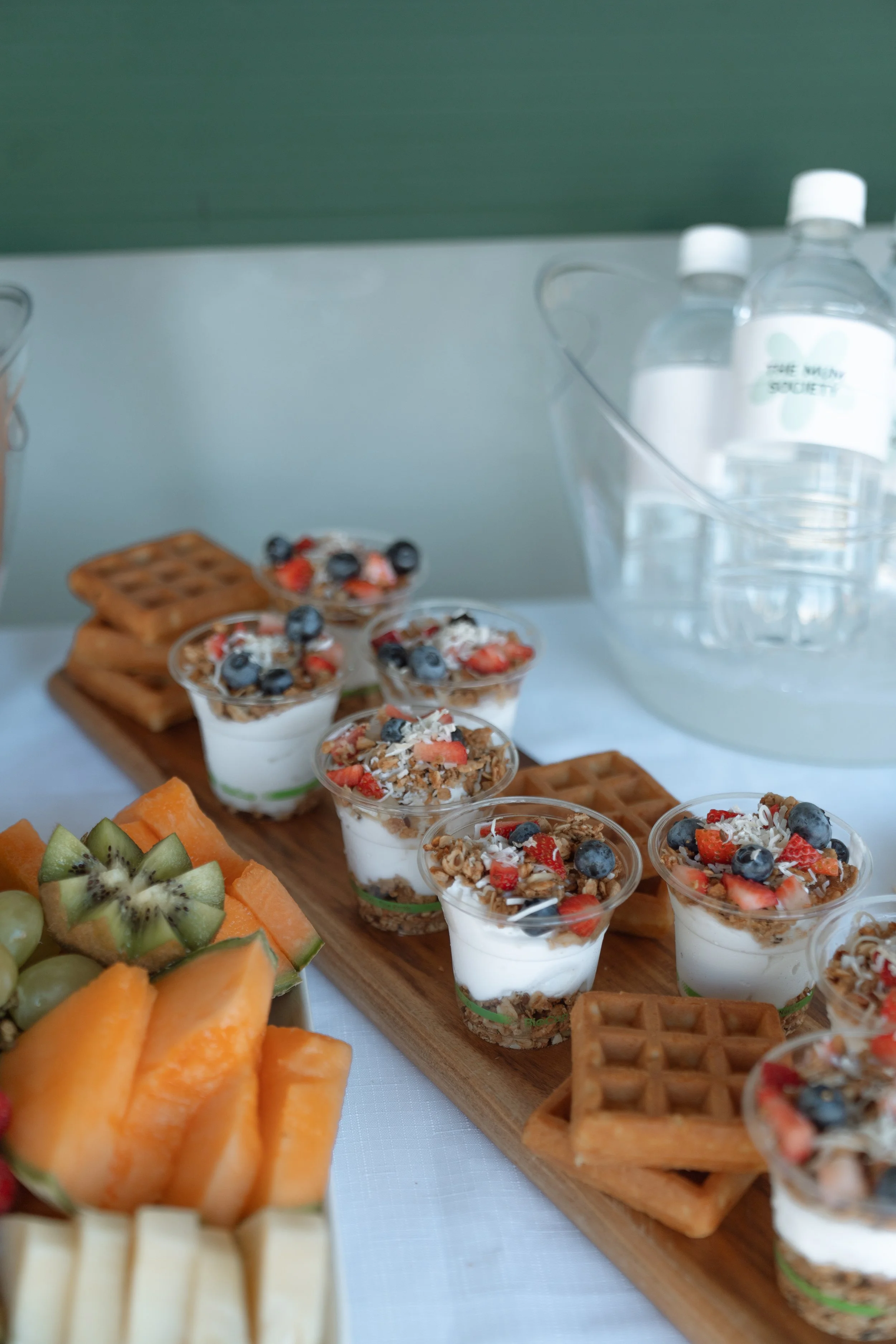 Tray of dessert parfaits topped with berries and coconut, alongside waffle pieces and fresh fruit on a table.