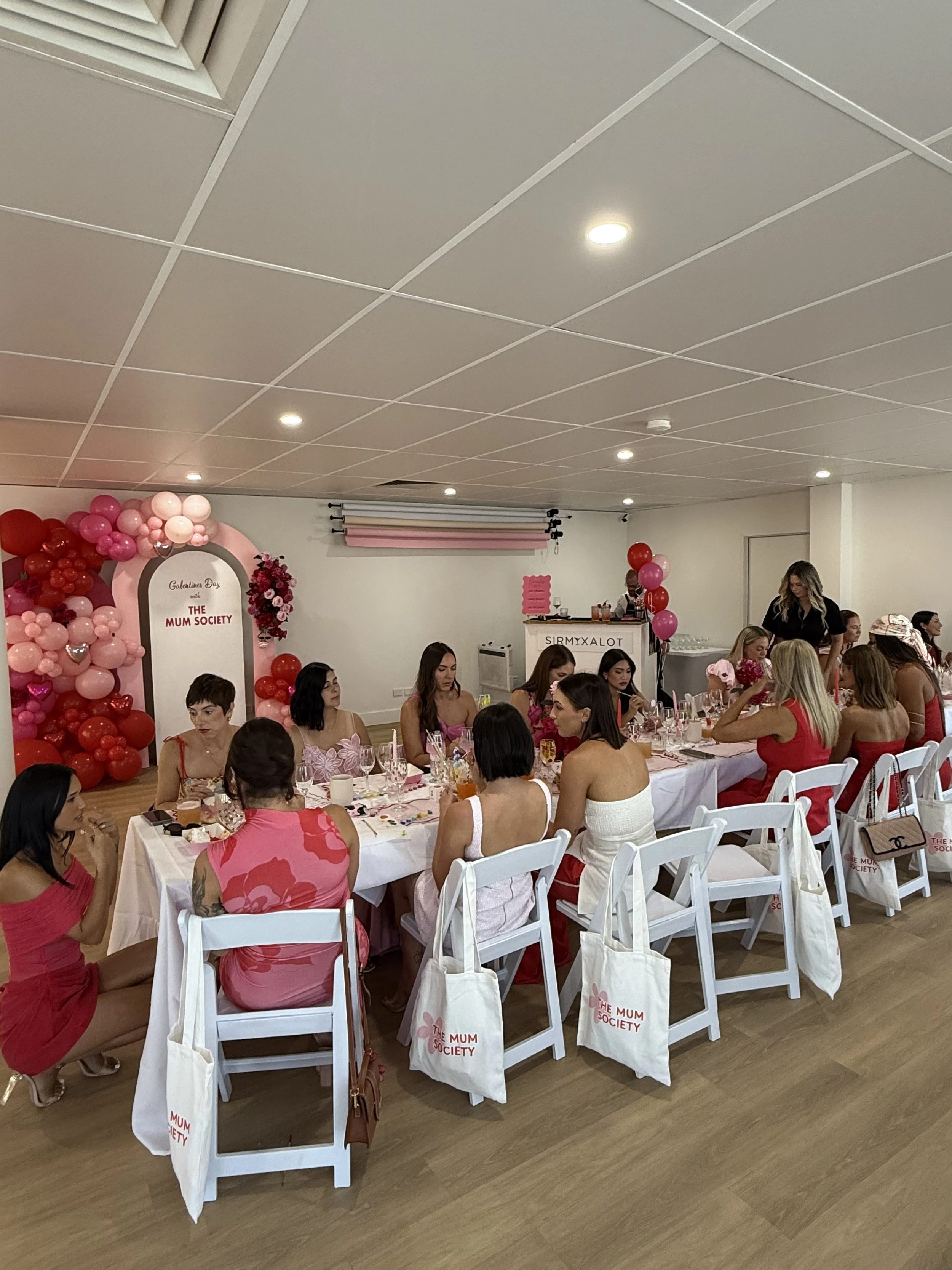 A gathering of women at a celebration event with pink and red decorations and balloons, sitting at a long table with food and drinks in a decorated room.