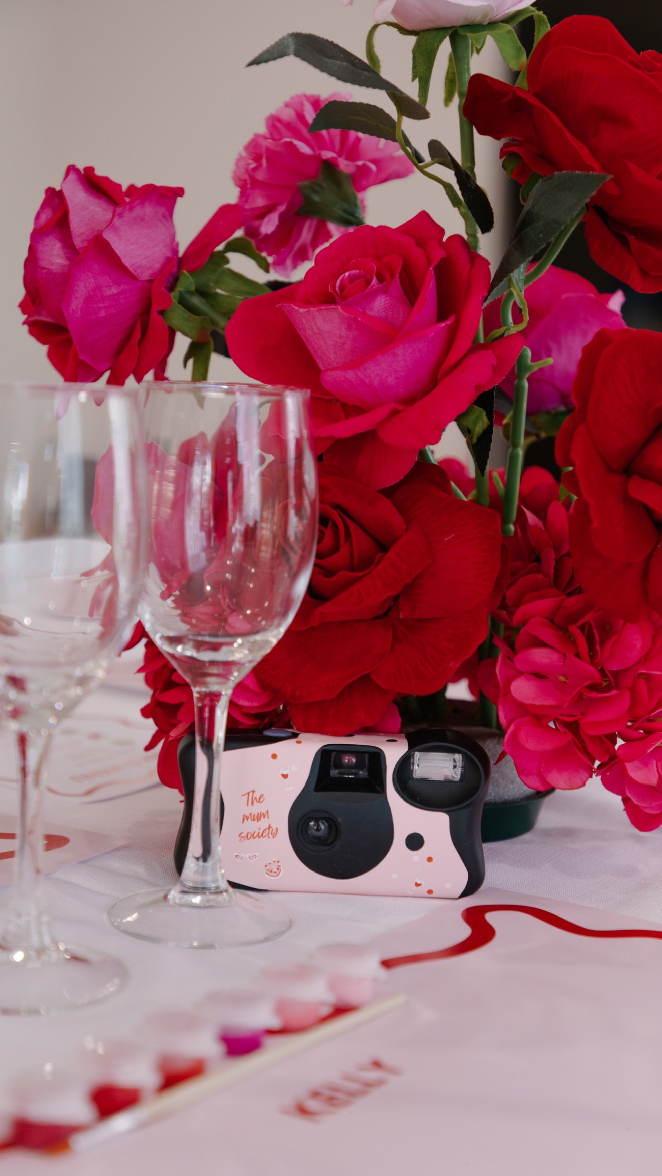 Pink and red roses in a bouquet on a table with empty wine glasses and a small camera, Valentine's Day decorations, and a tablecloth.