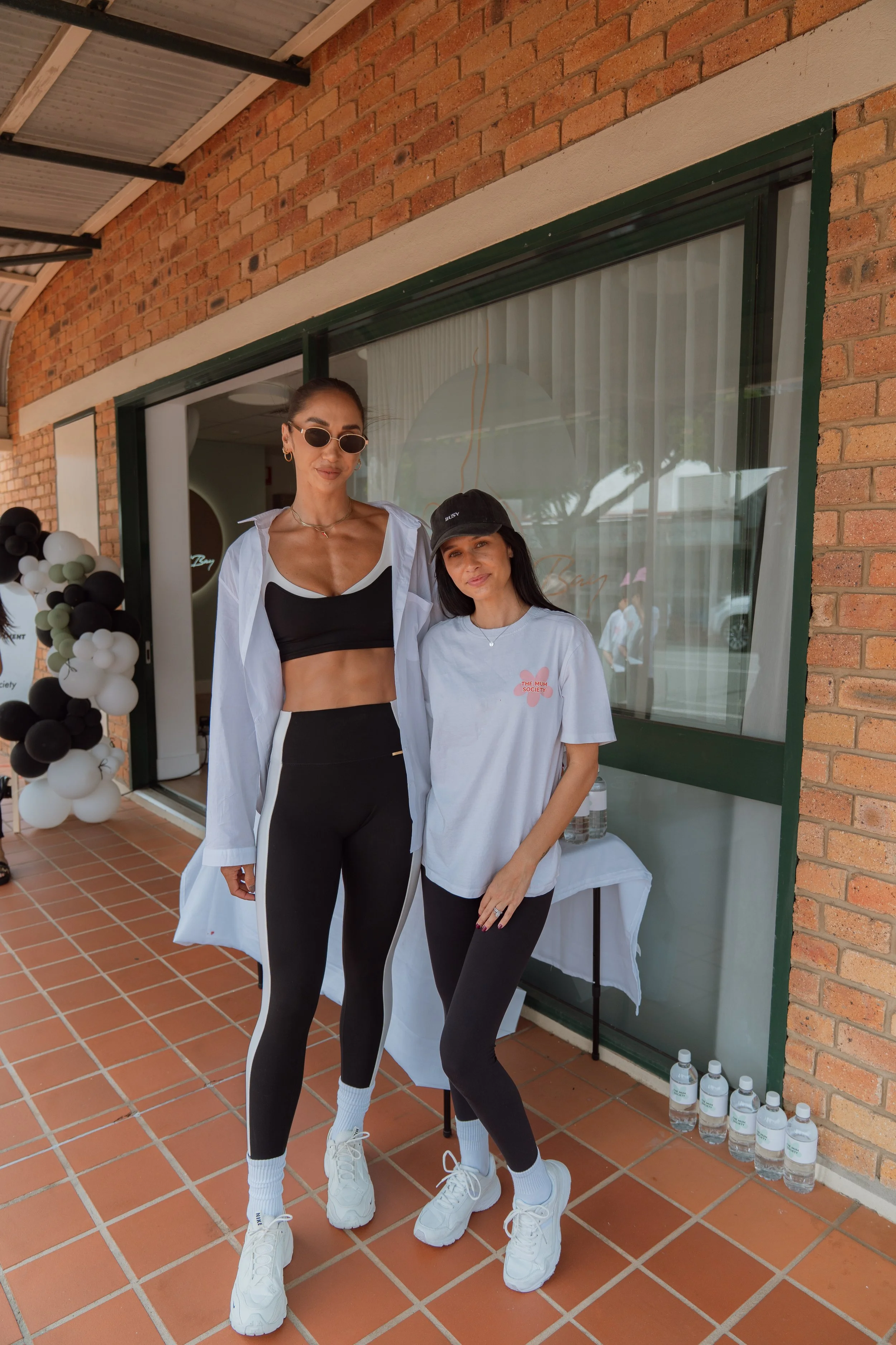 Two women posing in front of a brick building, with a table of water bottles behind them and a display of black, white, and green balloons to the side. One woman is tall, wearing black and white athletic clothing, white sneakers, and sunglasses. The 