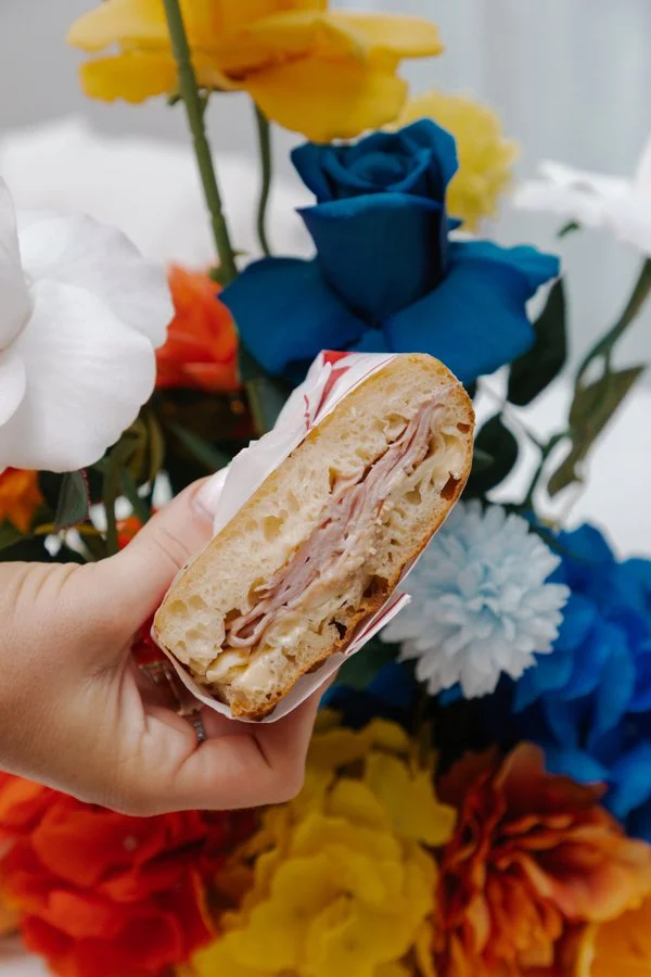 A hand holding a sandwich with deli meat, cheese, and bread in front of a colorful background of artificial flowers in yellow, blue, white, red, and orange.