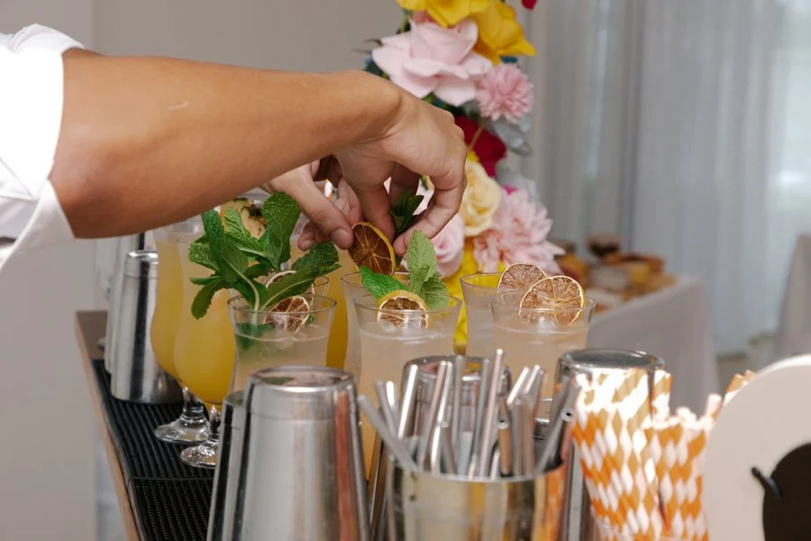 Person adding mint and dried lemon slices to glasses of cocktails or mocktails at a bar or refreshment table during a celebration, with a floral backdrop.