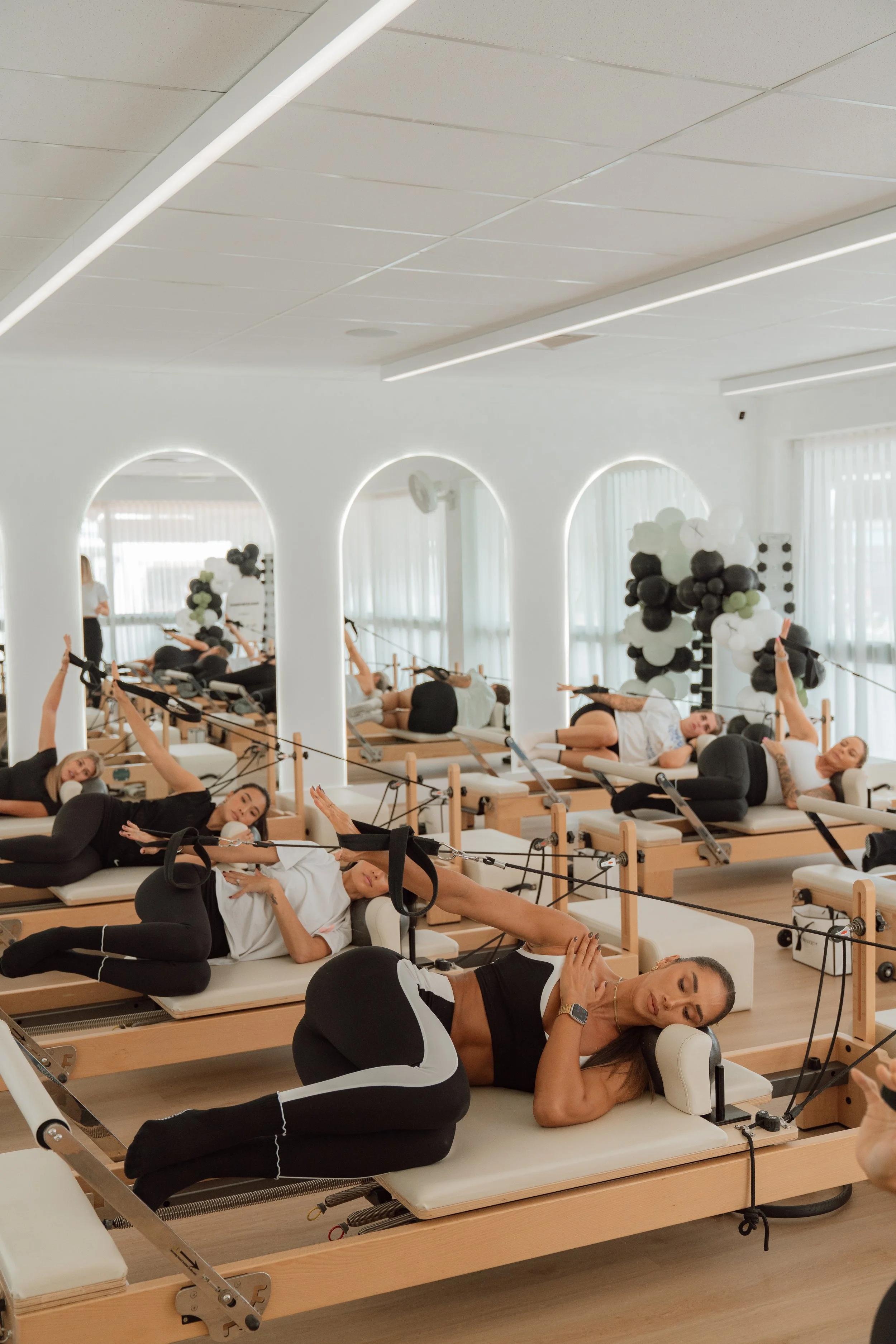 A Pilates class in a bright studio with mirrored walls, where women are lying on reformer machines performing stretches and exercises.