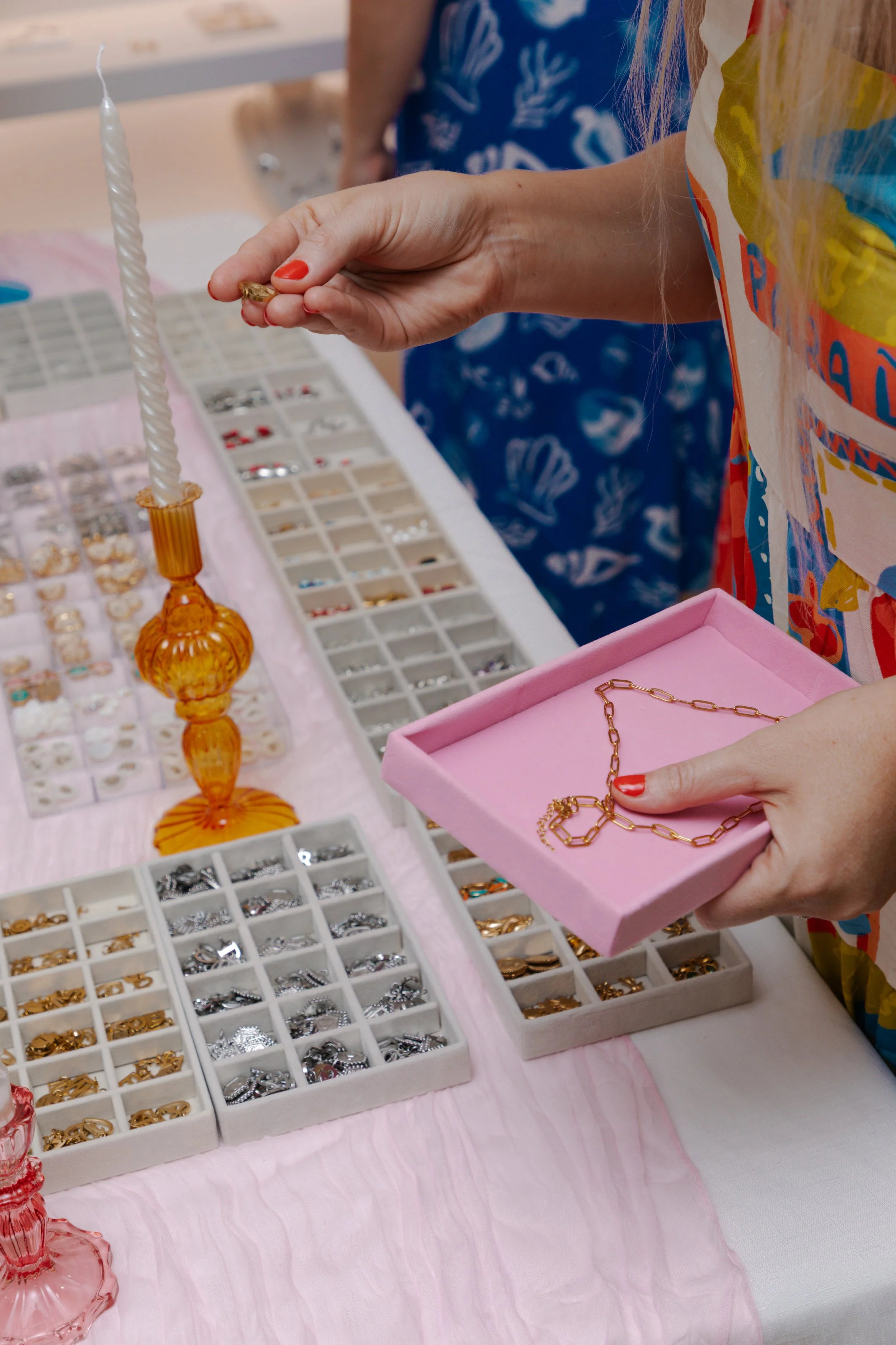 Person holding a pink jewelry tray with a gold chain necklace, surrounded by various jewelry items in display trays on a pink tablecloth.