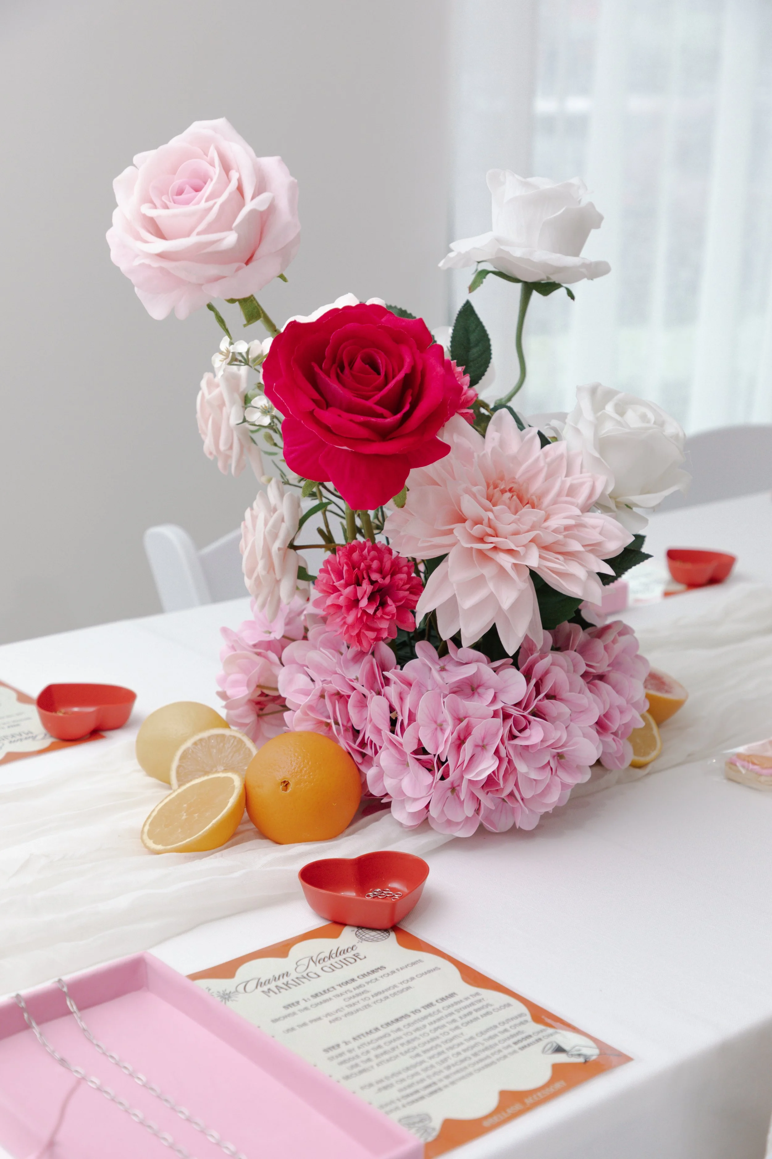 A floral centerpiece with pink, white, and red roses, and pink hydrangeas on a table with lemons, lemon slices, heart-shaped dishes, and a necklace-making guide.