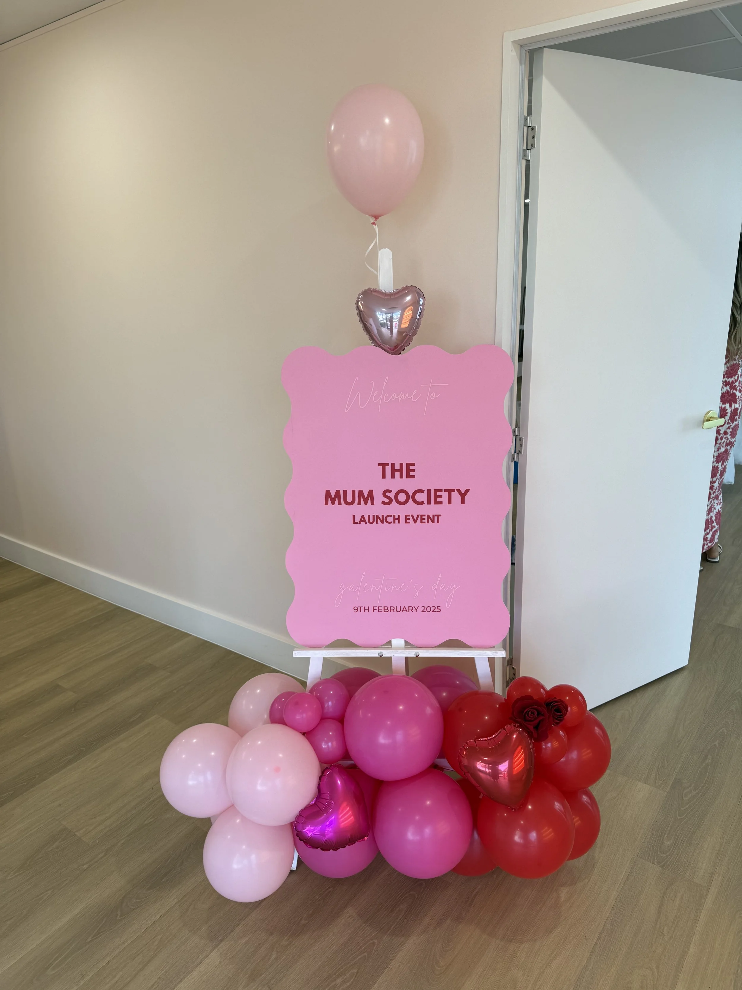 Decorative display for the Mum Society launch event, featuring pink and red balloons, heart-shaped balloons, and a pink welcoming sign with event details.