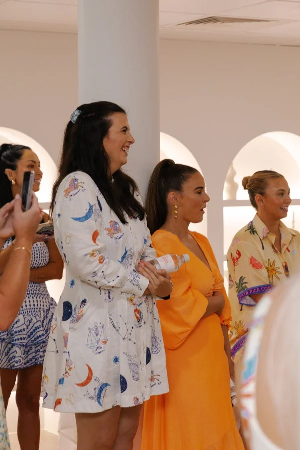 Group of women standing indoors, smiling, and watching an event or presentation. One woman is taking a photo with a smartphone. The women are dressed in colorful, patterned dresses.