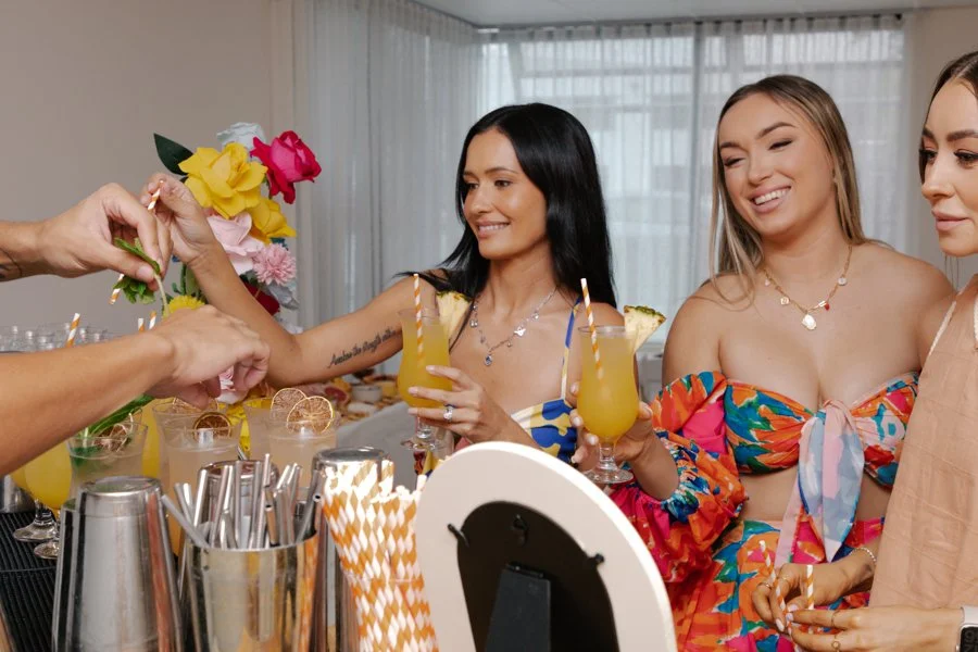 Three women in colorful dresses enjoying tropical drinks at a buffet, being served flowers.