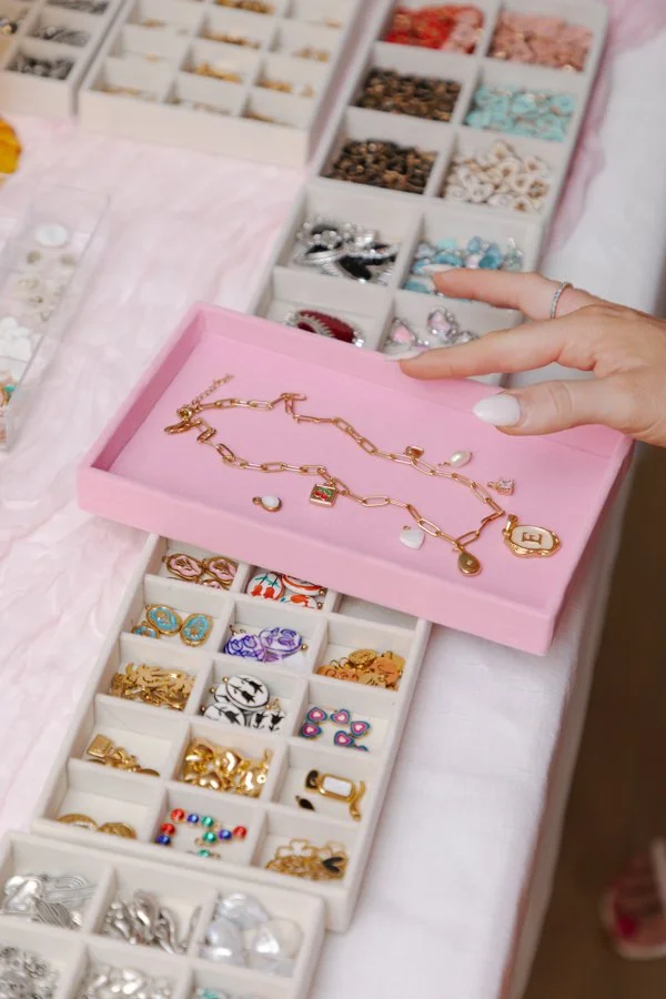 A person pointing to a pink jewelry box with a gold necklace inside, surrounded by various small jewelry charms and trinkets in organized compartments.
