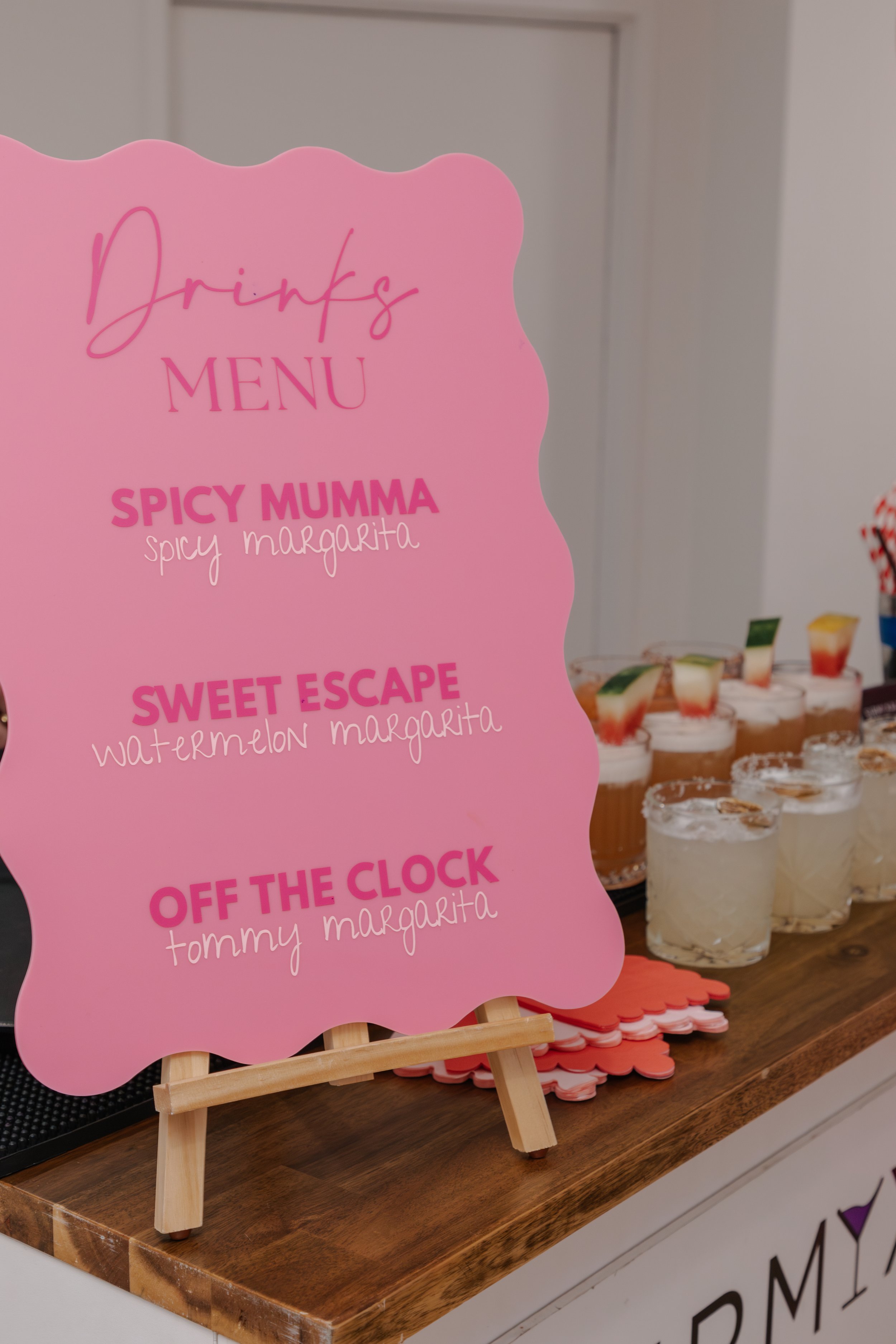 Pink drinks menu sign listing three beverages: Spicy Mumma (spicy margarita), Sweet Escape (watermelon margarita), Off the Clock (Tommy margarita). In the background, several glasses of decorated drinks are visible.