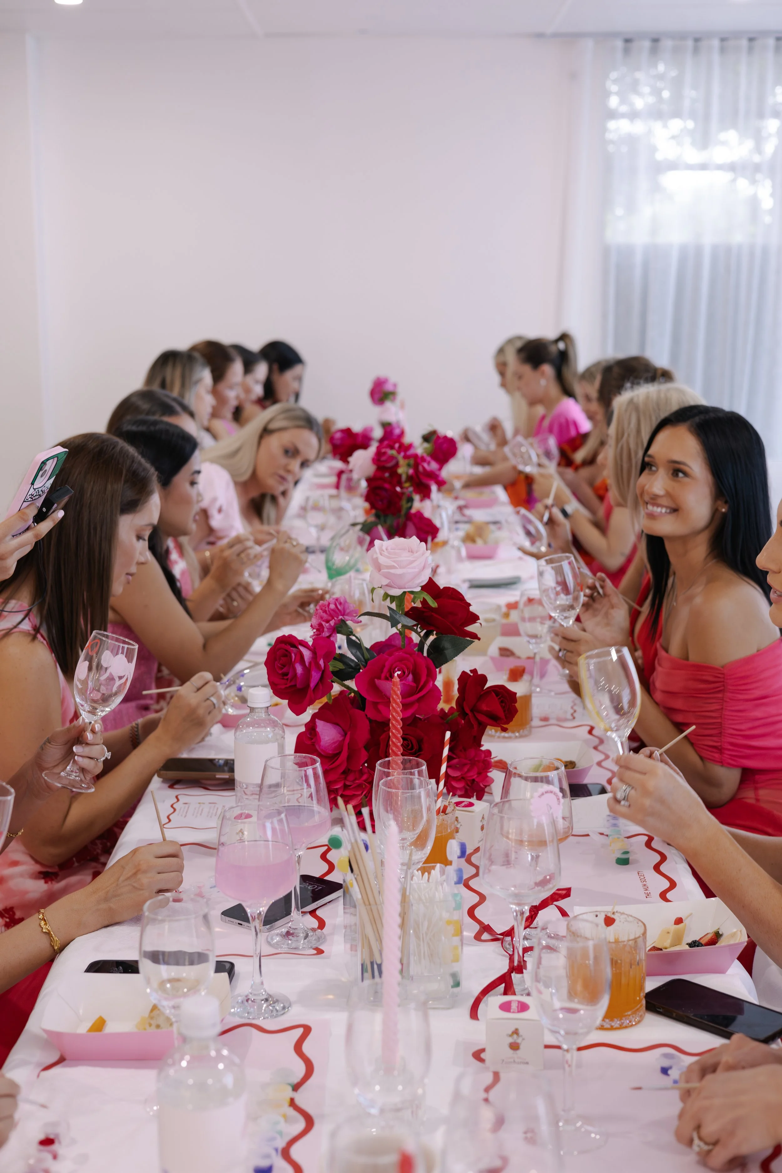 Women gathered around a decorated long table with pink and white flowers, wearing pink and red dresses, celebrating at a party or bridal shower.