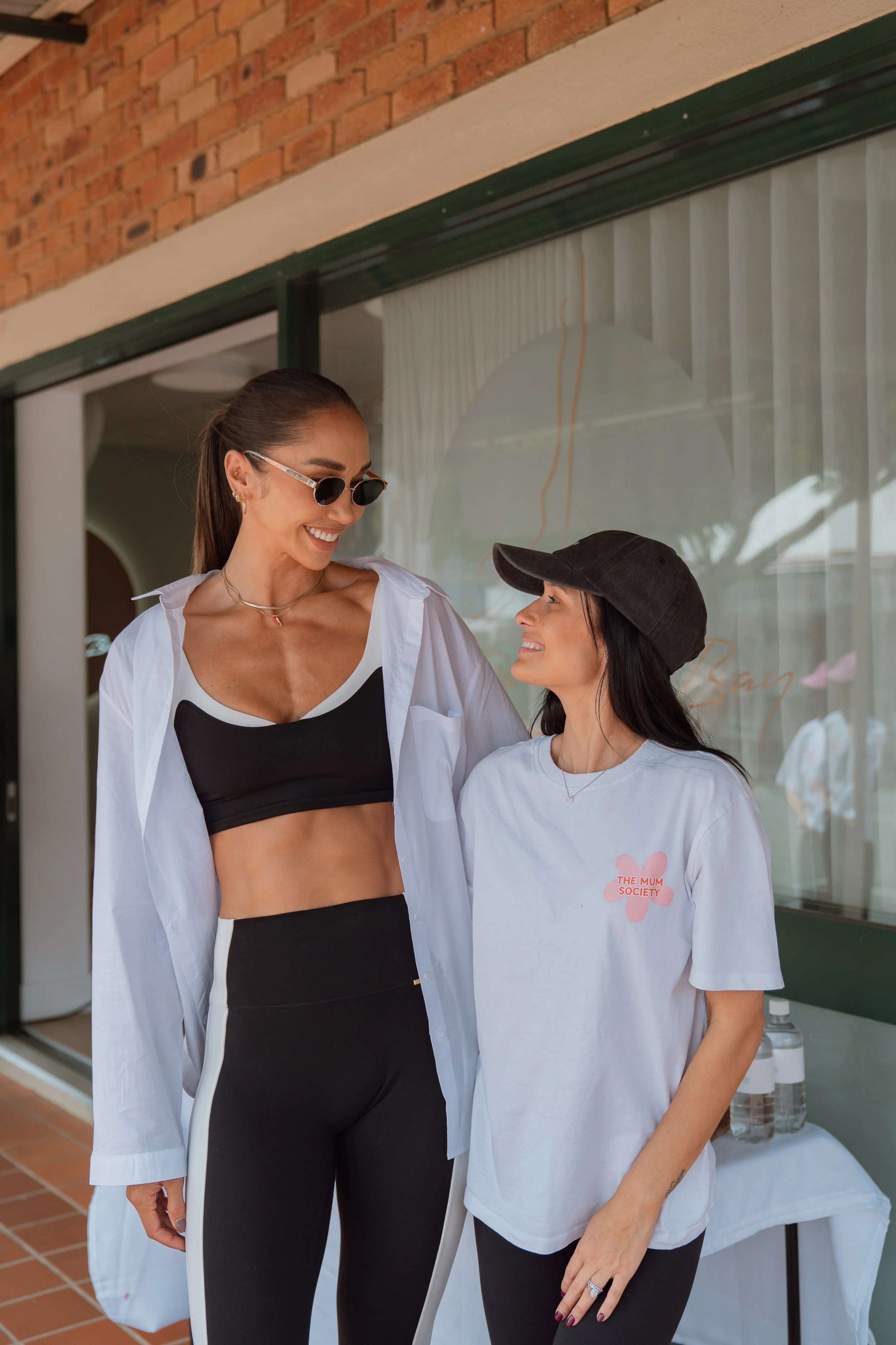 Two women standing close together, smiling and looking at each other, outdoors in front of a window with white curtains, brick wall, and glass door. One woman has long dark hair, wearing a black cap and a white T-shirt. The other woman has long brown
