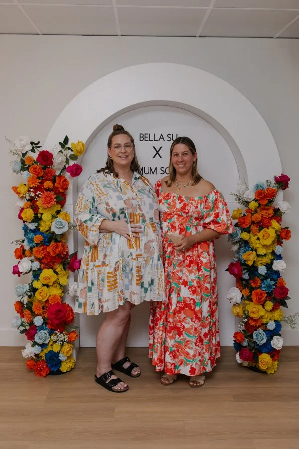 Two women standing in front of a floral arch and a sign that reads 'BELLA SULLIVAN X NUNU STORE'. They are smiling, wearing colorful dresses, and are in an indoor setting.