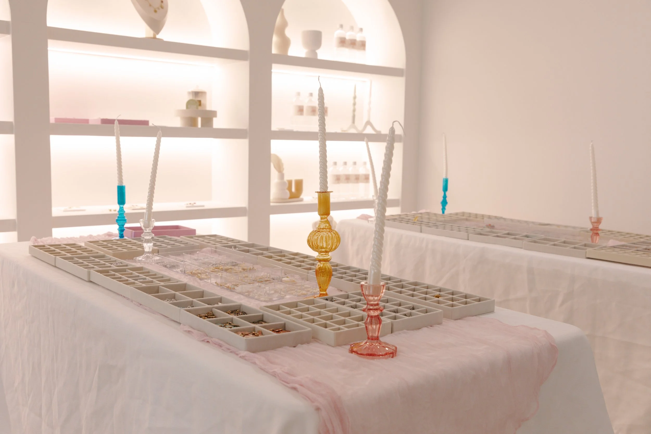 Tables set with jewelry display trays and tall, twisted candlesticks in transparent, blue, pink, and yellow glass, inside a bright, minimalist room with white shelves in the background.